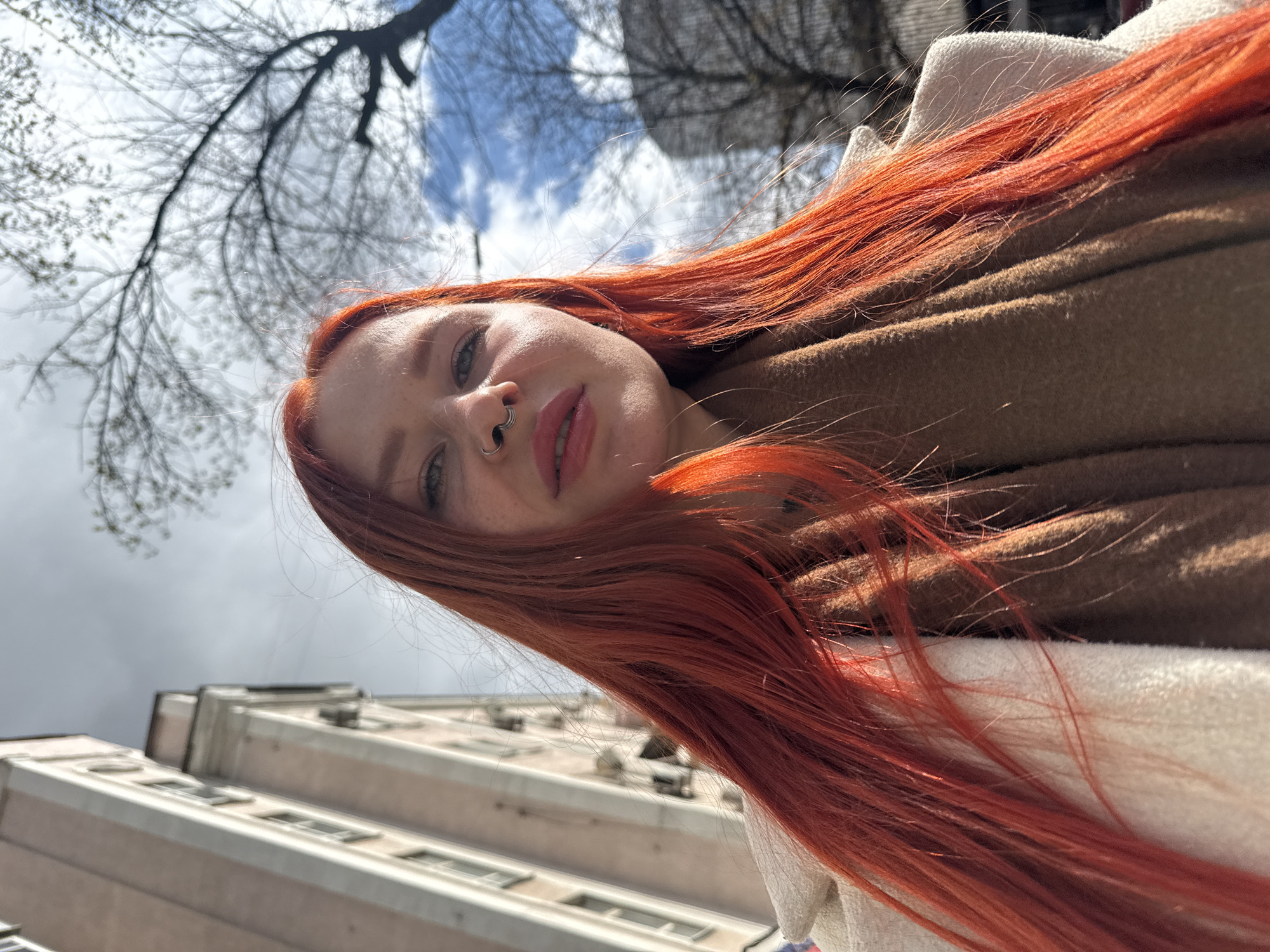 A low-angle selfie captured in Rostov-on-Don, Russia features a fair-skinned person with fiery red hair framing their face. They have freckles, a pierced nose, and are wearing pink lipstick. The person is wrapped in a beige scarf or garment, and a cream-colored jacket is visible. The backdrop includes a tall, light-colored building with multiple windows and what appear to be ventilation units or other architectural elements on its roof. Bare tree branches reach into a sky with patches of blue and gray clouds. The image appears to be taken during the day, with sunlight illuminating parts of the scene.