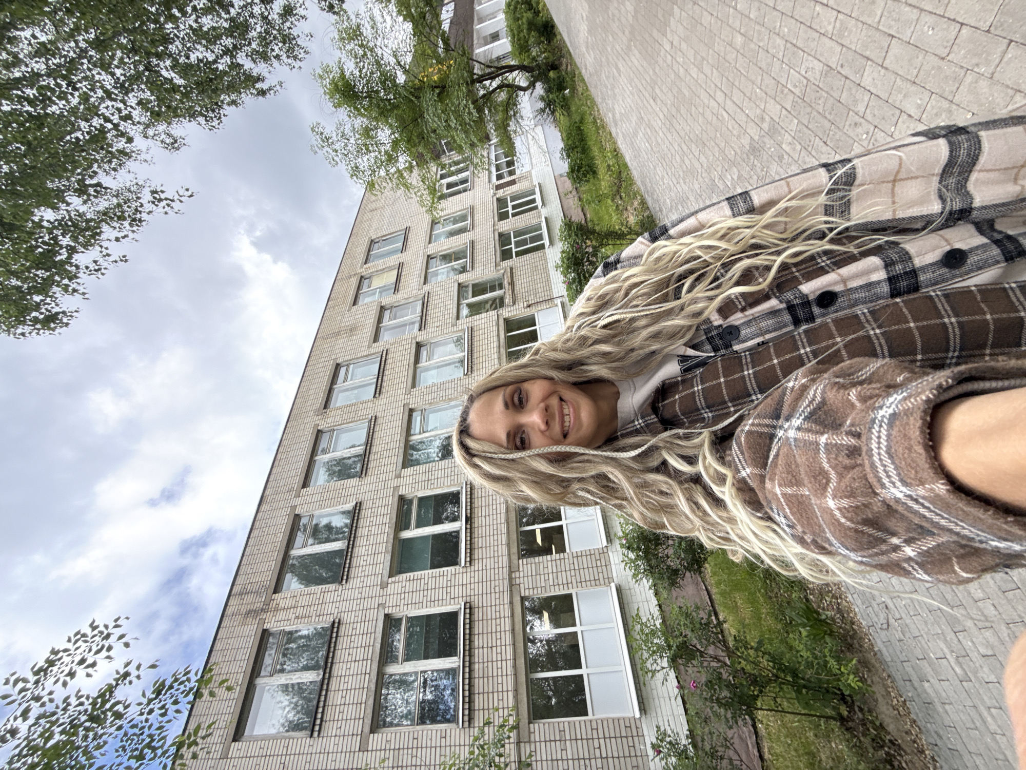 A woman poses in front of a brick building. She has long, blonde hair and is wearing a plaid shirt. The building has multiple windows. Trees are visible in the background, along with a sidewalk. The location is Rusanovo, Russia.