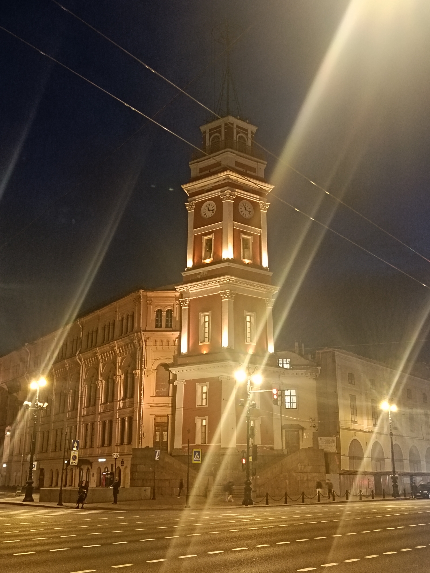 The image depicts a nighttime view of the Saint Petersburg Naval Institute in Russia. The building, with its distinctive tower featuring clocks on each side, is illuminated by warm lighting, contrasting with the dark blue sky. Streetlights cast bright rays, creating a halo effect. Pedestrians can be seen on the sidewalks near crosswalks. The street is lined with white lane markers. Overhead wires are visible against the sky.