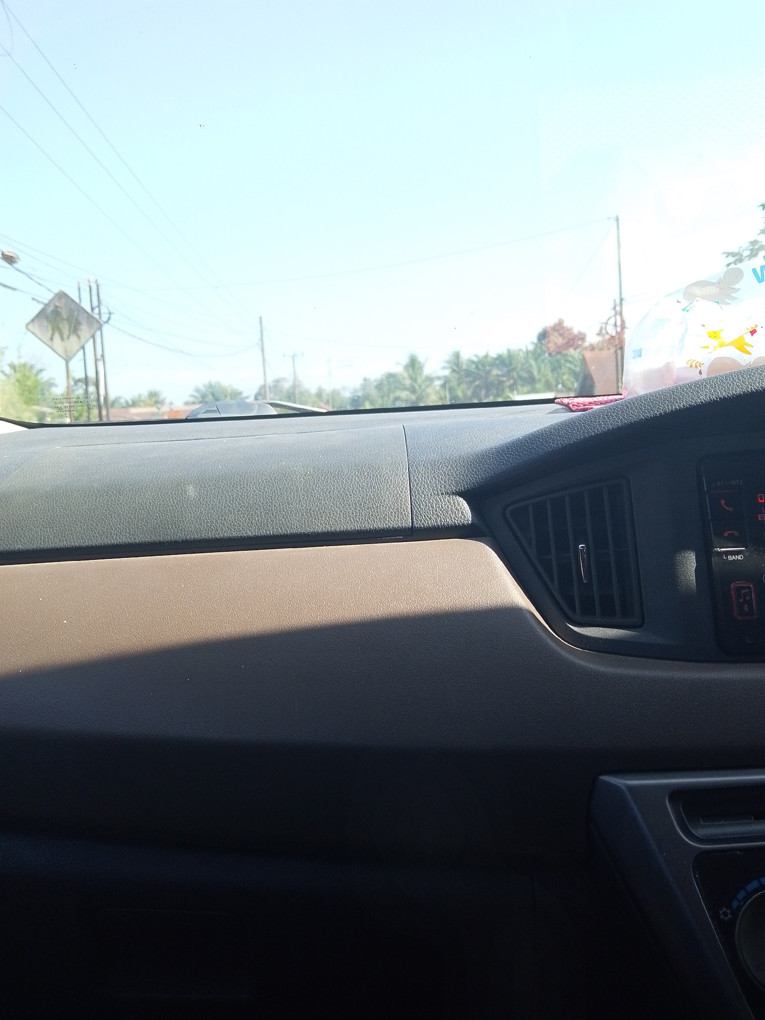 The image shows the interior of a car, looking out the windshield at a sunny, outdoor scene. The dashboard is visible in the foreground, with a textured black surface on the upper portion and a brown textured surface below. A rectangular air vent is visible on the right side of the dashboard. Outside the car, a clear blue sky is visible, with a few power lines strung across it. A diamond-shaped road sign depicting two figures walking is visible on the left side, along with trees and buildings in the distance. The scene appears to be in a semi-rural or suburban area. No people are directly visible within the car or interacting with the scene outside. The location is specified as Tiang Tara, Indonesia.