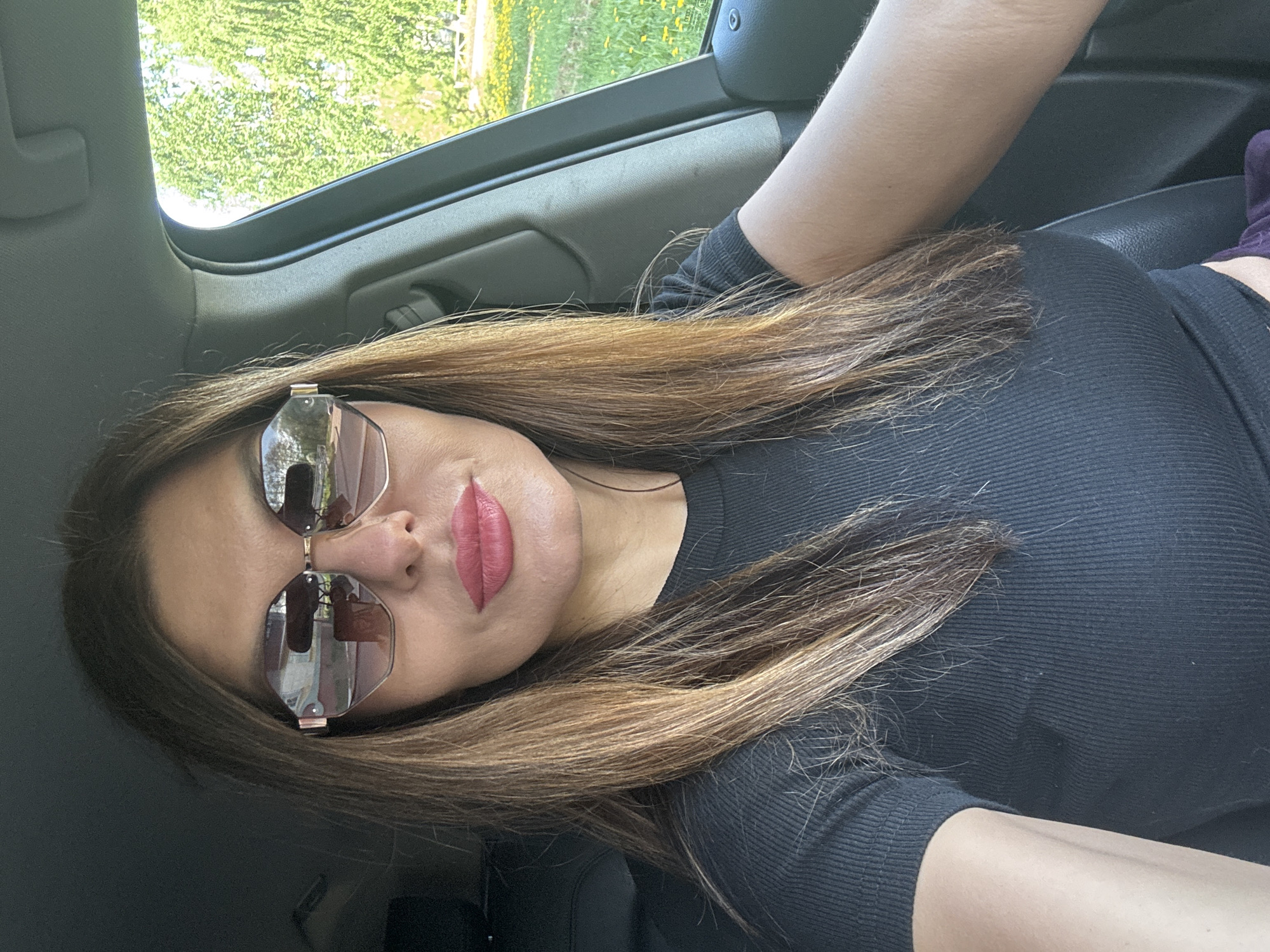 A woman is in a car, wearing sunglasses and a black shirt. The view out the car window shows greenery. It is not possible to determine if the location is Chaikovsky, Russia from this image alone.