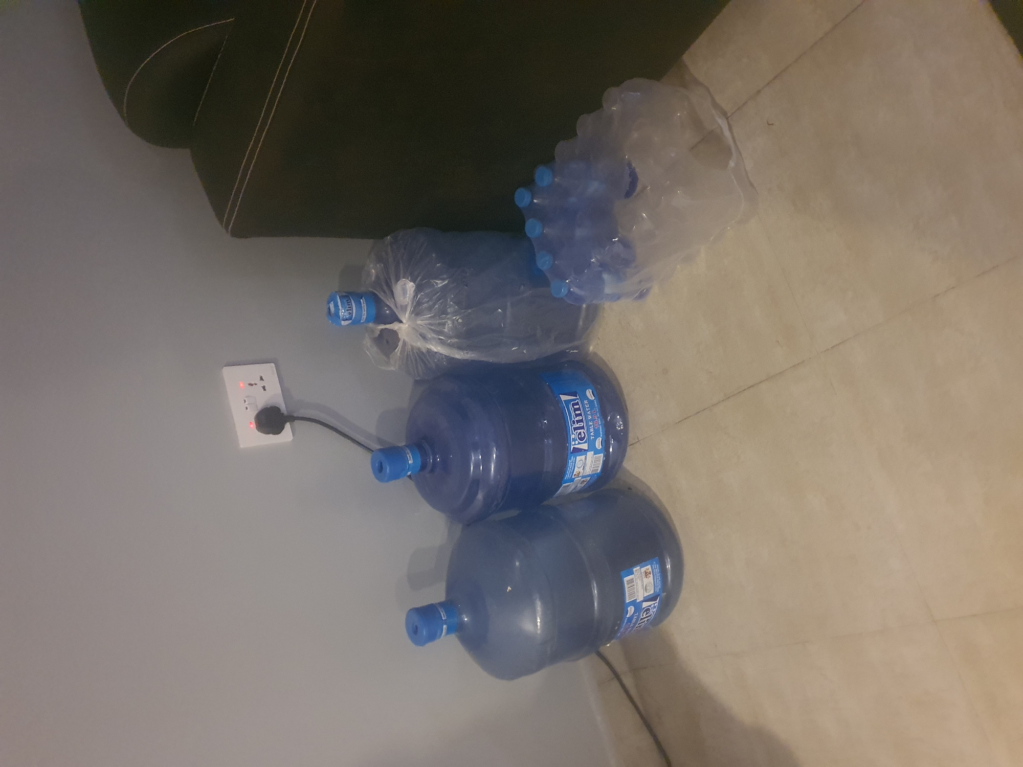 The image shows a collection of water bottles and large water jugs, likely for domestic use, placed on the floor next to a wall and a sofa. In the background, a portion of a dark green sofa with light-colored piping is visible. The foreground consists of a light gray wall and a tiled floor in a light beige color.
The primary subjects are several large, clear plastic water jugs with blue caps. Two of these jugs are clearly visible, stacked one on top of the other. They both have blue labels with white text that appears to read "Elim Table Water". One of the jugs has a white sticker with some writing on it. In addition to the large jugs, there are also several smaller plastic water bottles bundled together in a clear plastic bag, with blue caps visible. Another large, clear plastic bag contains a single large water jug, also with a blue cap and a visible label.
To the left of the water jugs, a white electrical outlet is mounted on the gray wall. It has a black plug inserted into it, and a red indicator light is illuminated on the outlet. A black cord extends from the plug.
The scene appears to be indoors, given the presence of the sofa and the electrical outlet. The lighting suggests it might be daytime or a well-lit indoor space, but there are no clear indicators of the time of day or weather conditions. The overall impression is one of a residential setting where water is being stored, possibly due to a shortage or for convenience. There are no people visible in the image.