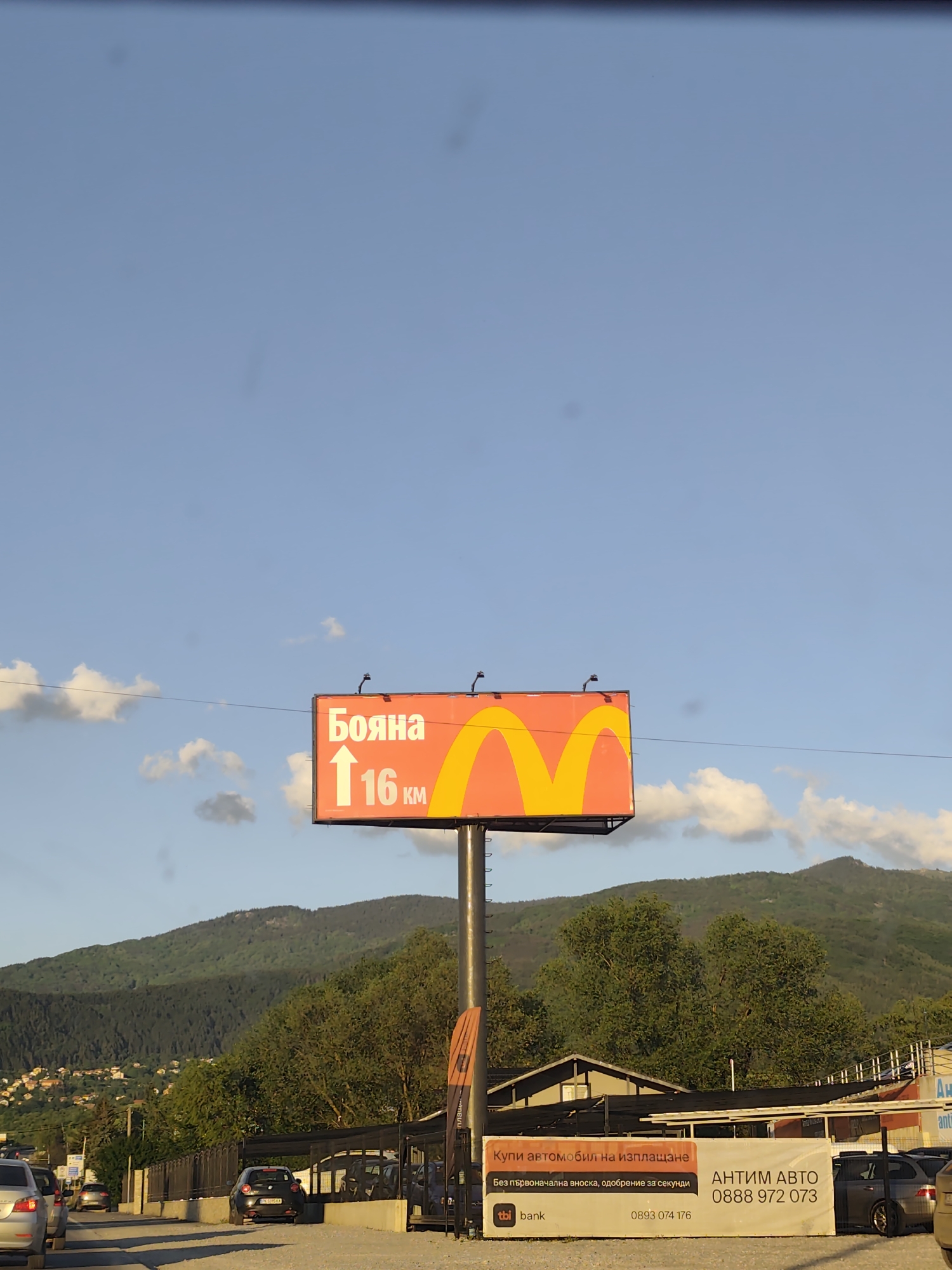 The image shows a McDonald's billboard in the city of Dragichevo, Bulgaria. The billboard features the McDonald's logo along with a sign indicating that Boyana is 16 kilometers away. Below the billboard, there is a sign advertising "Antim Avto" and TBI Bank, promoting car purchases with financing options. In the background, there are mountains, trees, and some buildings. Cars are parked in front of the businesses, and there is traffic on the road. The sky is blue with scattered clouds.