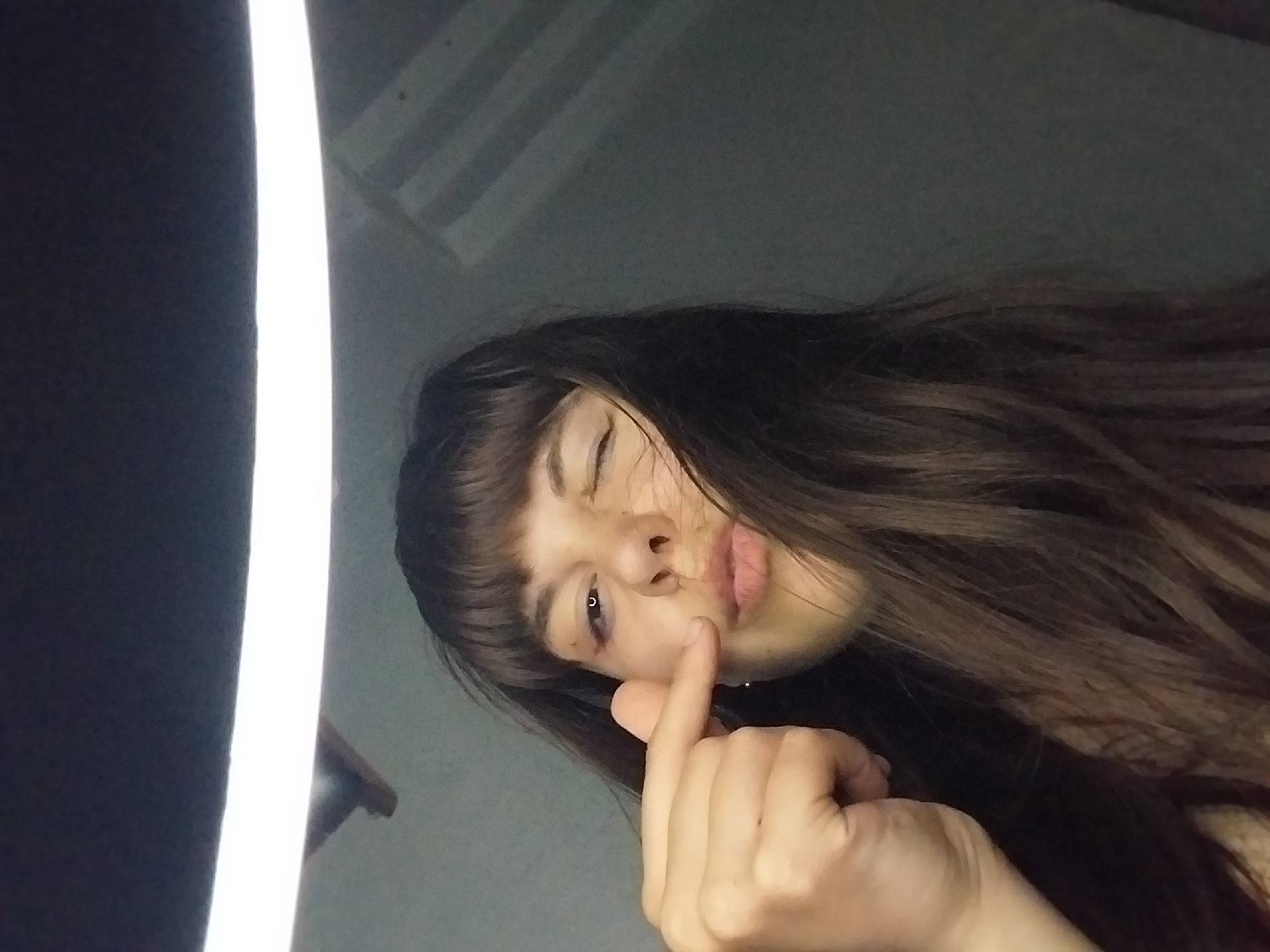A young woman with dark hair and bangs is looking directly at the camera, winking with her left eye. Her right eye is open, revealing a slight purple hue near the eyelid, suggesting a possible bruise or shadow. She has her left hand raised, with her index finger extended and pointing towards her cheekbone. Her lips are slightly parted. The lighting in the image is dramatic, with a bright ring light illuminating her face from above, casting shadows on the background, which appears to be a textured, grey wall. The overall impression is a selfie taken in an indoor setting.