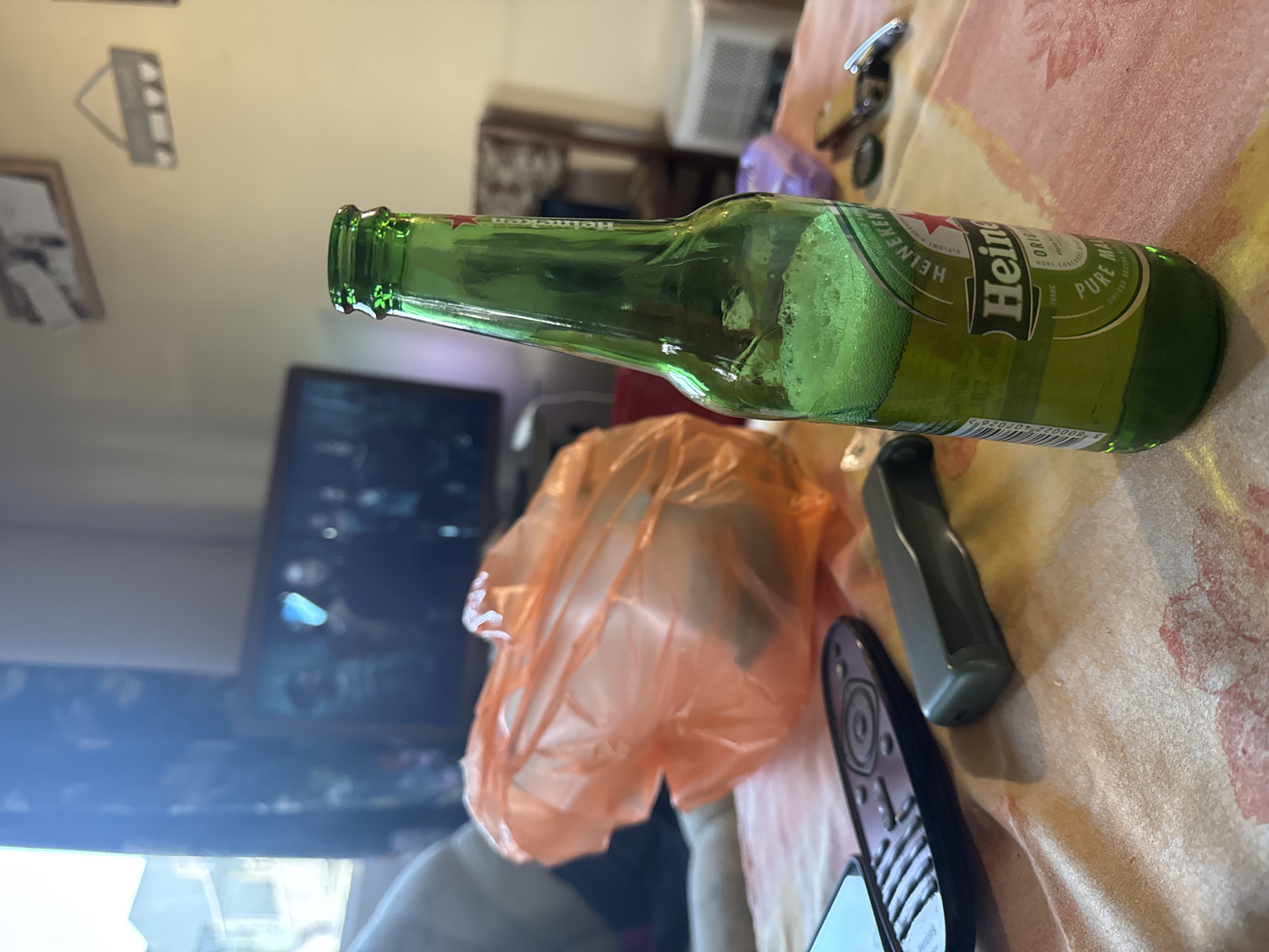 The image shows a partially empty green glass bottle of Heineken beer lying horizontally on a surface with a patterned tablecloth. An orange plastic bag sits next to the bottle, along with two TV remotes. In the background, a TV screen is visible, along with other household items. The location is unknown.
