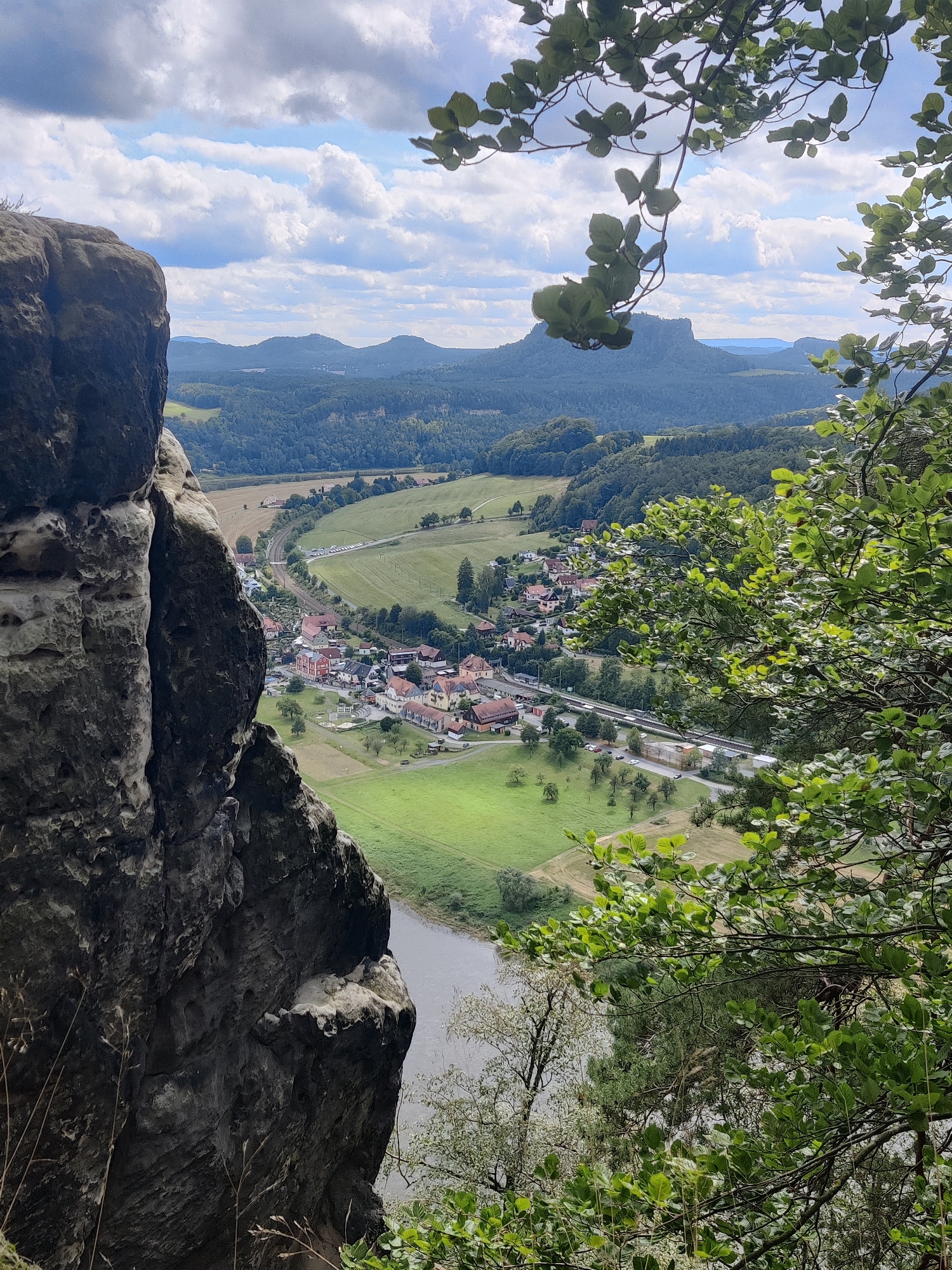 This is a photograph taken from a high vantage point in Lohmen, Germany, looking down on a village nestled in a valley. In the foreground, a large, rugged rock formation juts out from the left side of the frame, its rough texture and dark color dominating the immediate view. Branches of lush green foliage hang down from the upper right, partially obscuring the view of the landscape beyond.
Below the rock and foliage, a village is visible, characterized by a cluster of buildings with red-tiled roofs, interspersed with smaller structures and green spaces. A winding road or railway line runs through the village, with several cars parked along its edge. Surrounding the village are expanses of vibrant green fields and agricultural land, divided by patches of trees.
In the middle distance, the landscape rises into rolling hills covered in dense green forests. Further in the background, a distinctive mesa-like rock formation stands out against the horizon, a prominent geological feature. The sky above is a mix of blue with scattered white clouds. A body of water, likely a river, can be seen flowing through the valley near the village. The overall impression is one of a scenic, rural landscape with a visible settlement integrated into the natural environment.