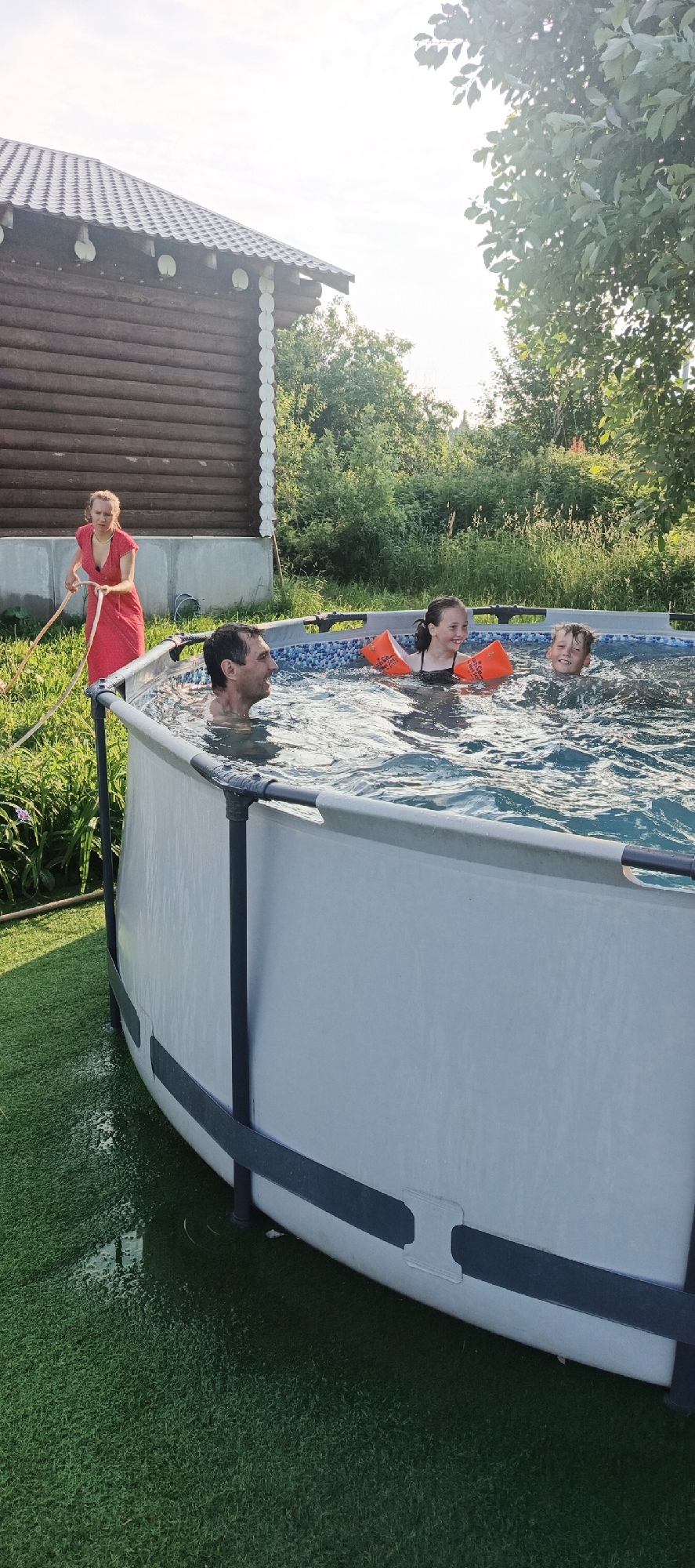 In the village of Bolshoi Bukor, Russia, an above-ground pool is set up on a grassy area next to a wooden house. A woman in a red dress is holding a hose, appearing to be filling or cleaning the pool. Inside the pool, a man and two children are swimming. The children are wearing orange arm floaties and are smiling. The scene suggests a warm, sunny day, likely during the summer, as indicated by the outdoor activity and the lush greenery. The setting appears to be a residential backyard.