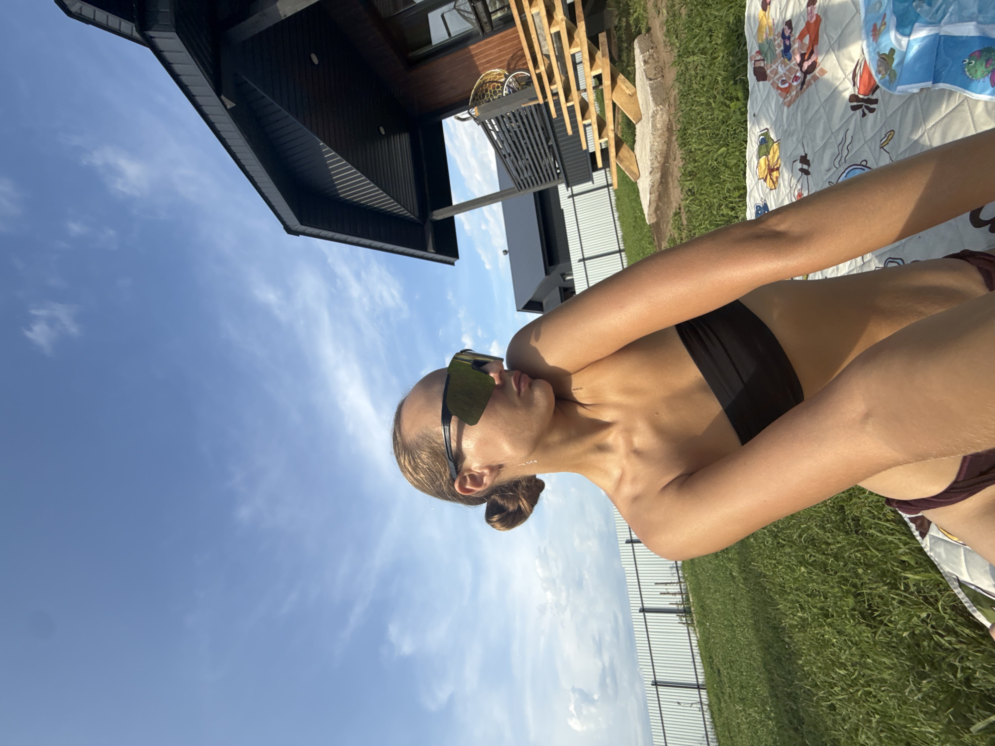 A young woman is captured in a selfie taken outdoors in a grassy area. She is wearing a black bandeau bikini top and dark bottoms. Large, dark sunglasses obscure her eyes, and her blonde hair is styled in a bun. Her left arm is bent and resting near her head, while her right arm is extended, holding the camera. She is positioned on a blanket with a playful pattern. In the background, a modern black house with wooden accents and stairs is visible, along with a white metal fence and a clear blue sky with scattered clouds. The scene suggests a warm, sunny day.