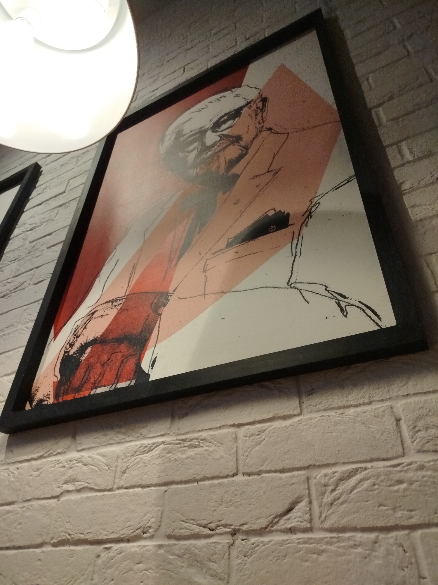 The image captures an indoor scene, primarily featuring a framed artwork mounted on a textured white brick wall. A large, bright white circular light fixture is partially visible in the upper left quadrant, casting illumination over the area.
The artwork is a contemporary, stylized illustration of a man, likely an older gentleman, depicted from the chest up. He is shown wearing glasses and a suit jacket with a visible pocket square. The artistic style combines bold black outlines with abstract, geometric blocks of red and peach-colored areas, set against a white background. The man's facial features are rendered with an expressive, almost sketchy quality. The picture is encased in a simple black frame.
The wall behind the artwork and light fixture is constructed from white or off-white bricks, displaying clear texture and visible mortar lines. The lighting from the fixture appears to be artificial, highlighting the rough surface of the bricks and creating subtle shadows. The photograph is taken from a low-angle perspective, looking upwards towards the art and light source. No discernible text is visible on the artwork itself. The setting evokes a modern interior design, potentially a commercial establishment such as a restaurant or cafe located in Varna, Bulgaria.