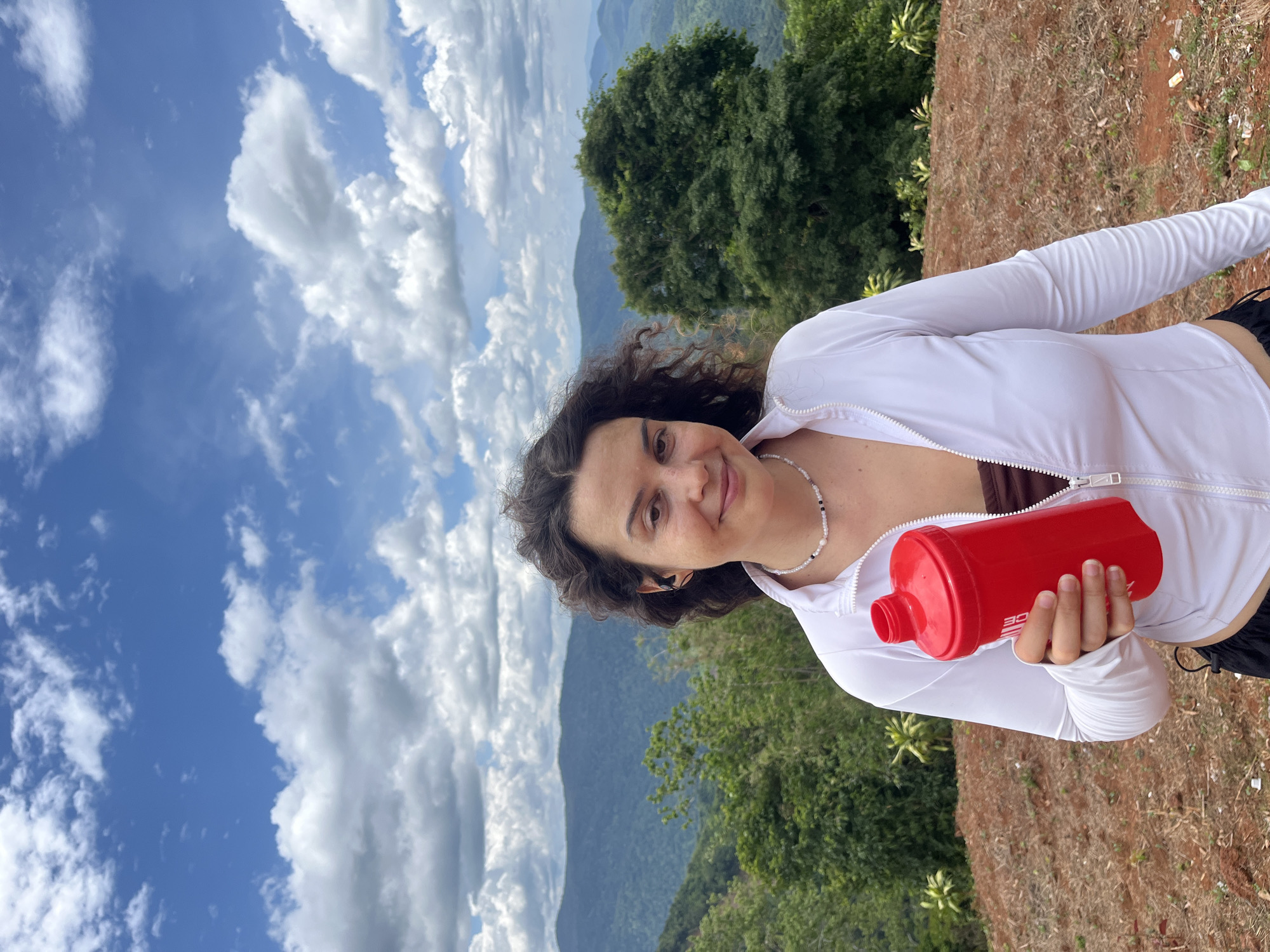 A young woman stands outdoors holding a red shaker bottle in her left hand. She is wearing a white zip-up jacket, a brown top, and black shorts. She has curly dark brown hair and is looking at the camera with a slight smile. The background consists of lush green mountains and a cloudy blue sky. The location is identified as Nao Se, Vietnam. The ground in the foreground is reddish-brown soil.