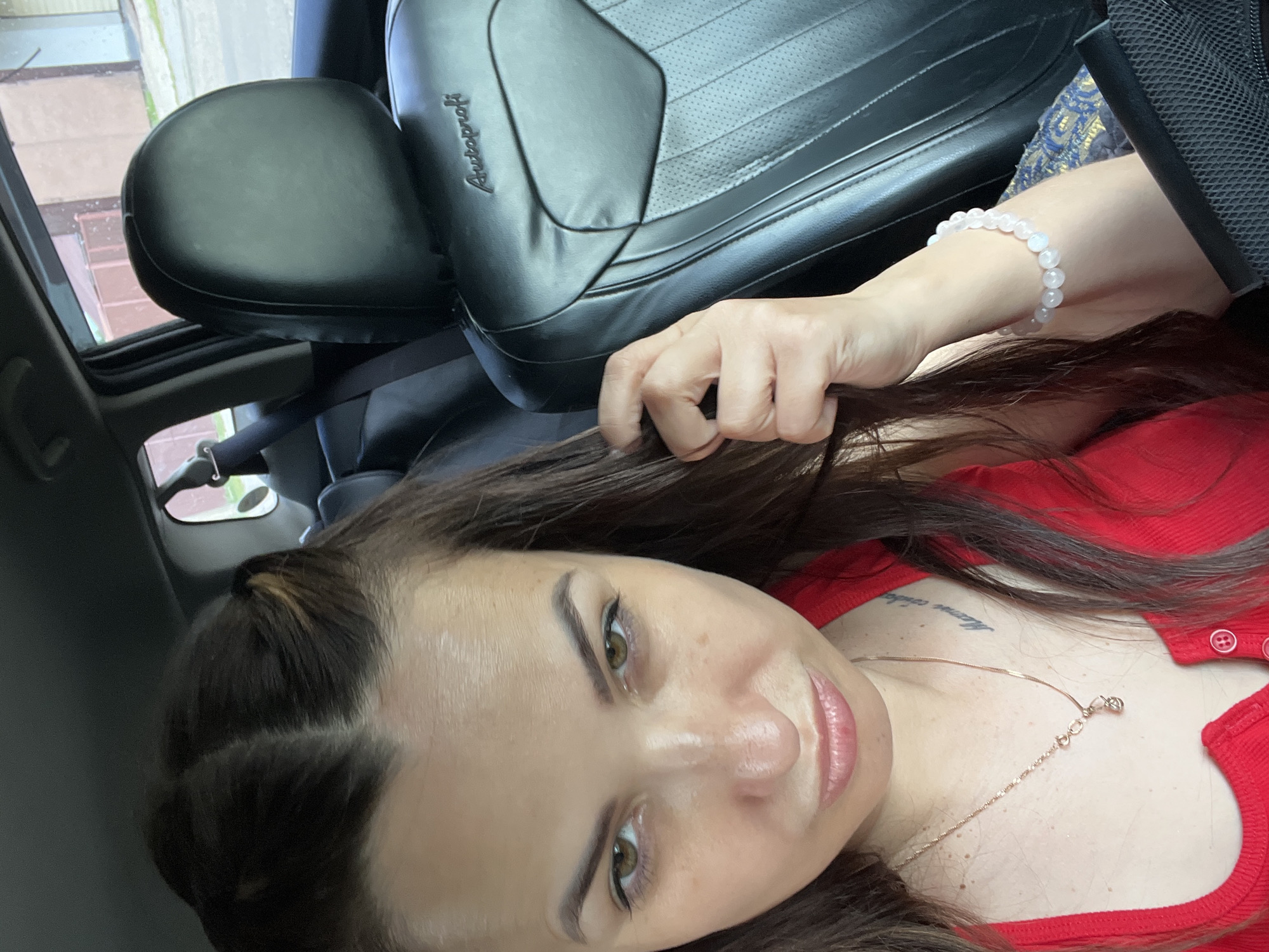 A woman is seated in the passenger seat of a car, looking at the camera. She has dark hair pulled back in a partial updo, and is wearing a red tank top. A rose gold necklace with a small pendant is visible around her neck. She has a bracelet of light pink beads on her left wrist. The car seat next to her is black leather with the brand name "Autoprofi" embossed on it. The background shows the interior of the car, with a window offering a view of buildings and trees outside. The scene appears to be taking place in a city, though the specific location is unknown.