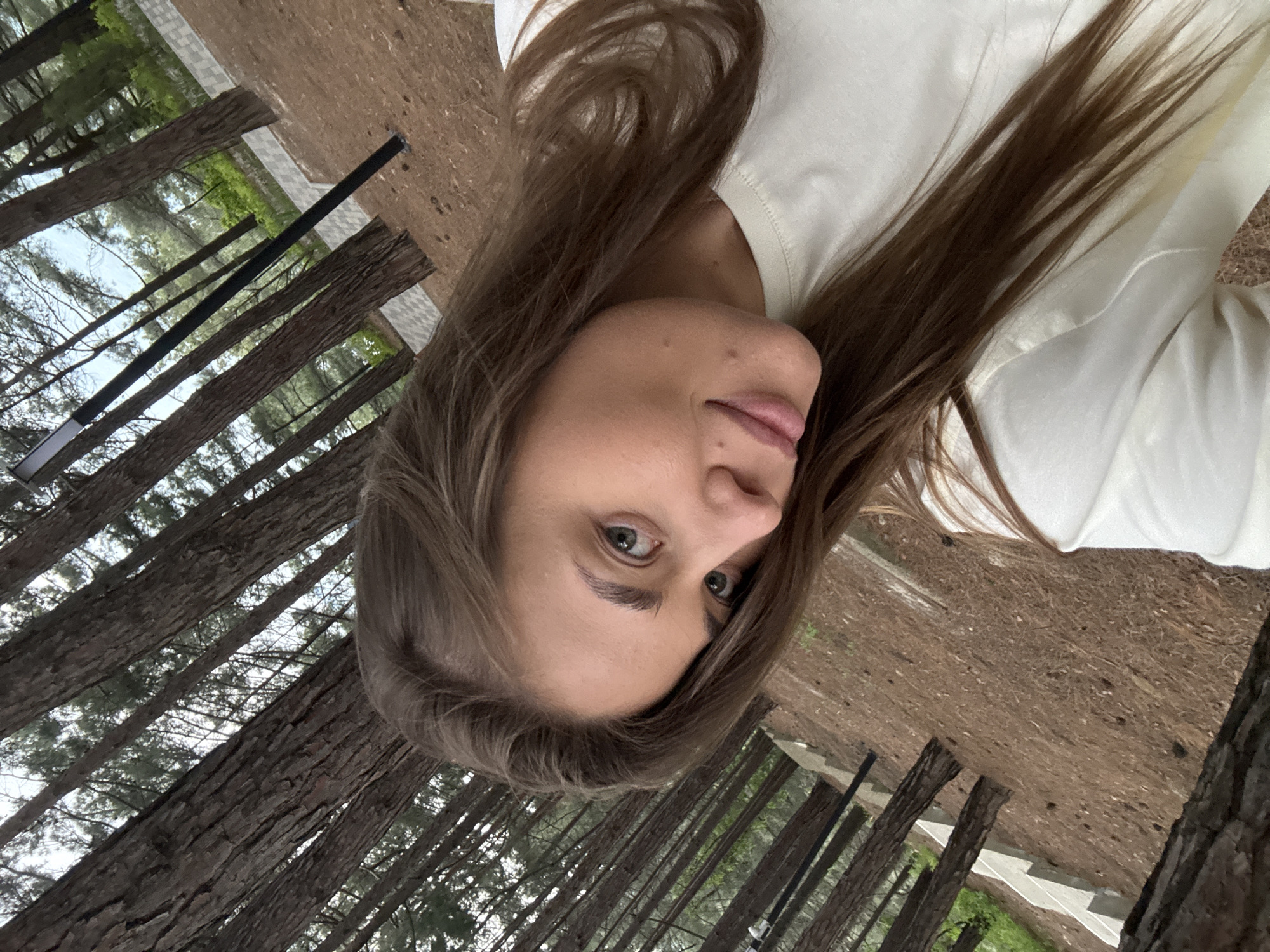 A woman with brown hair and blue eyes is the subject of the image. She is wearing a white top. The background appears to be a forest or wooded area, possibly within the city of Sosnovy, Russia. The trees are tall and slender. The image is taken at an unusual angle.