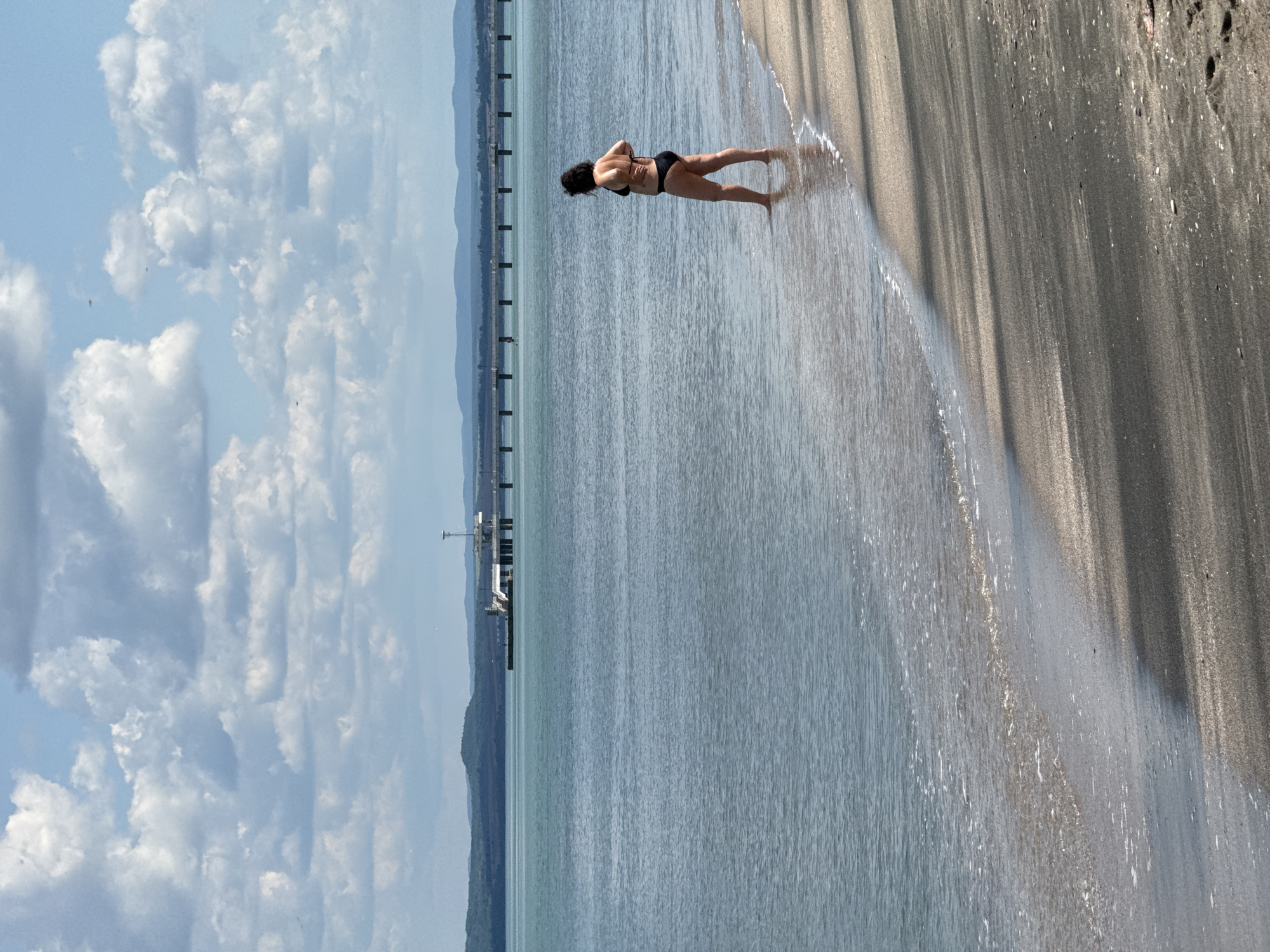 A woman in a black bikini stands in the shallows of a beach. The beach has dark sand and water. The sky is blue with puffy white clouds. In the distance is a long pier extending into the water and, further back, a mountain range. The location is Burgas, Bulgaria, as indicated by the Burgas Bridge visible in the distance.
