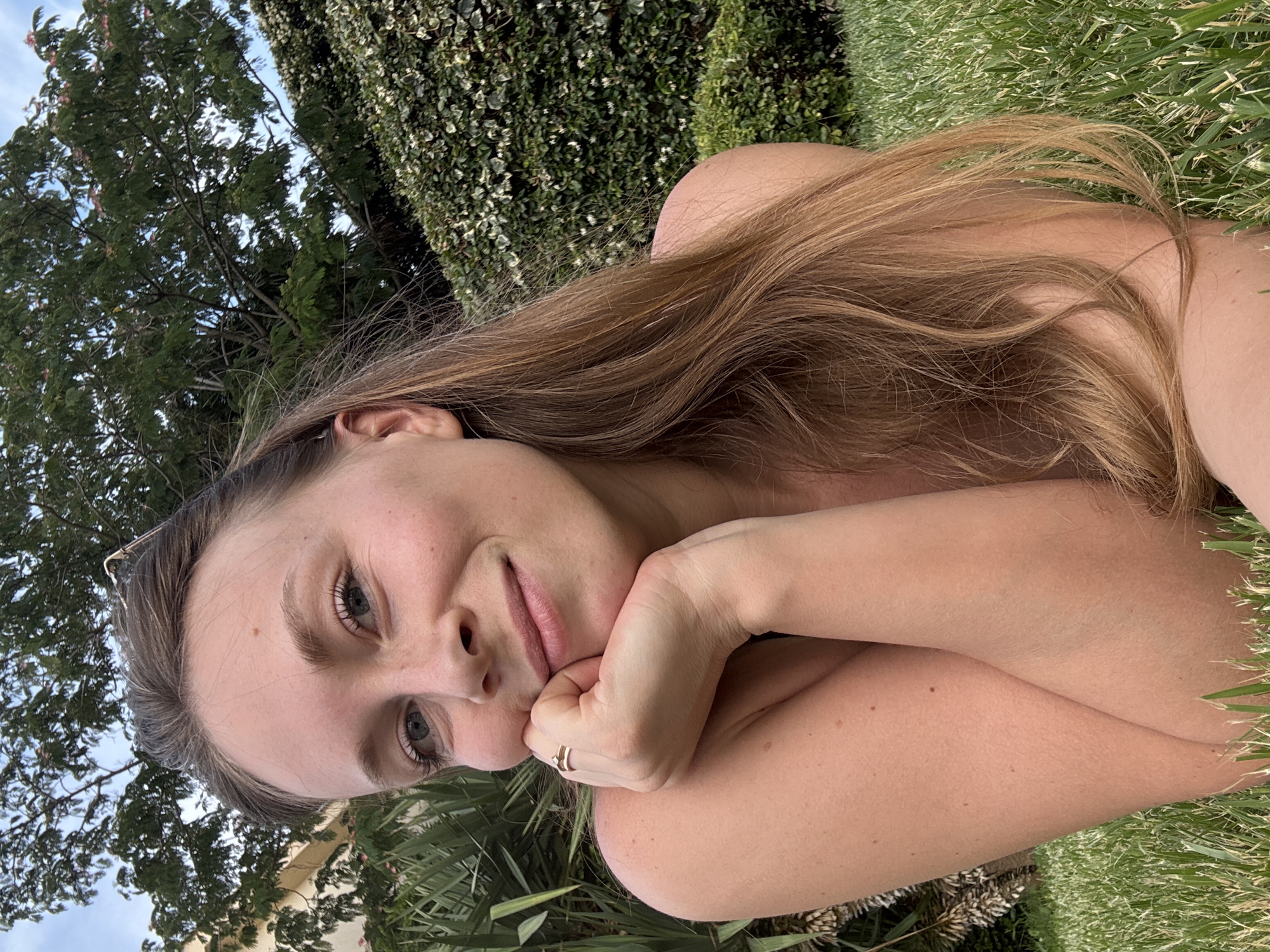 The media file shows a young woman lying on the grass, with a wall of ivy and trees in the background. She is looking towards the camera with a relaxed expression. Her hand is resting on her chin, and she is wearing a ring on her finger. Her long, light brown hair is spread out around her. The scene appears to be set outdoors in a garden or park, with plenty of greenery and natural light. The time of day seems to be daytime, possibly afternoon, given the brightness of the sunlight. The overall mood is peaceful and serene. There is no visible text in the media file. The location context is Sveti Stefan, Montenegro.