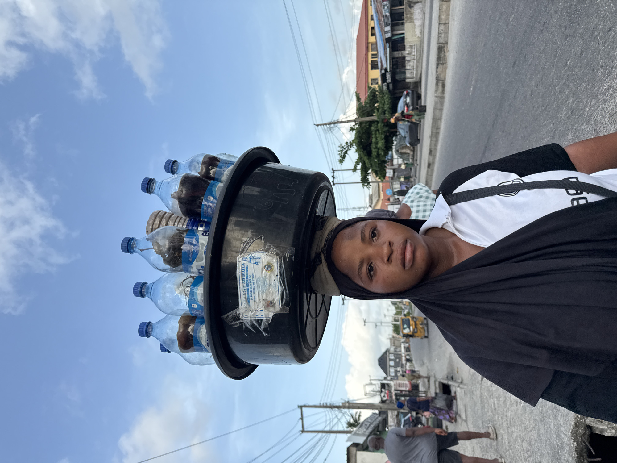 A woman balances a black bucket filled with water bottles on her head. The bucket has a label attached. She wears a black headscarf, a white t-shirt, and a black shawl. The background shows a street in Ado, Nigeria, with buildings, utility poles, and other people.