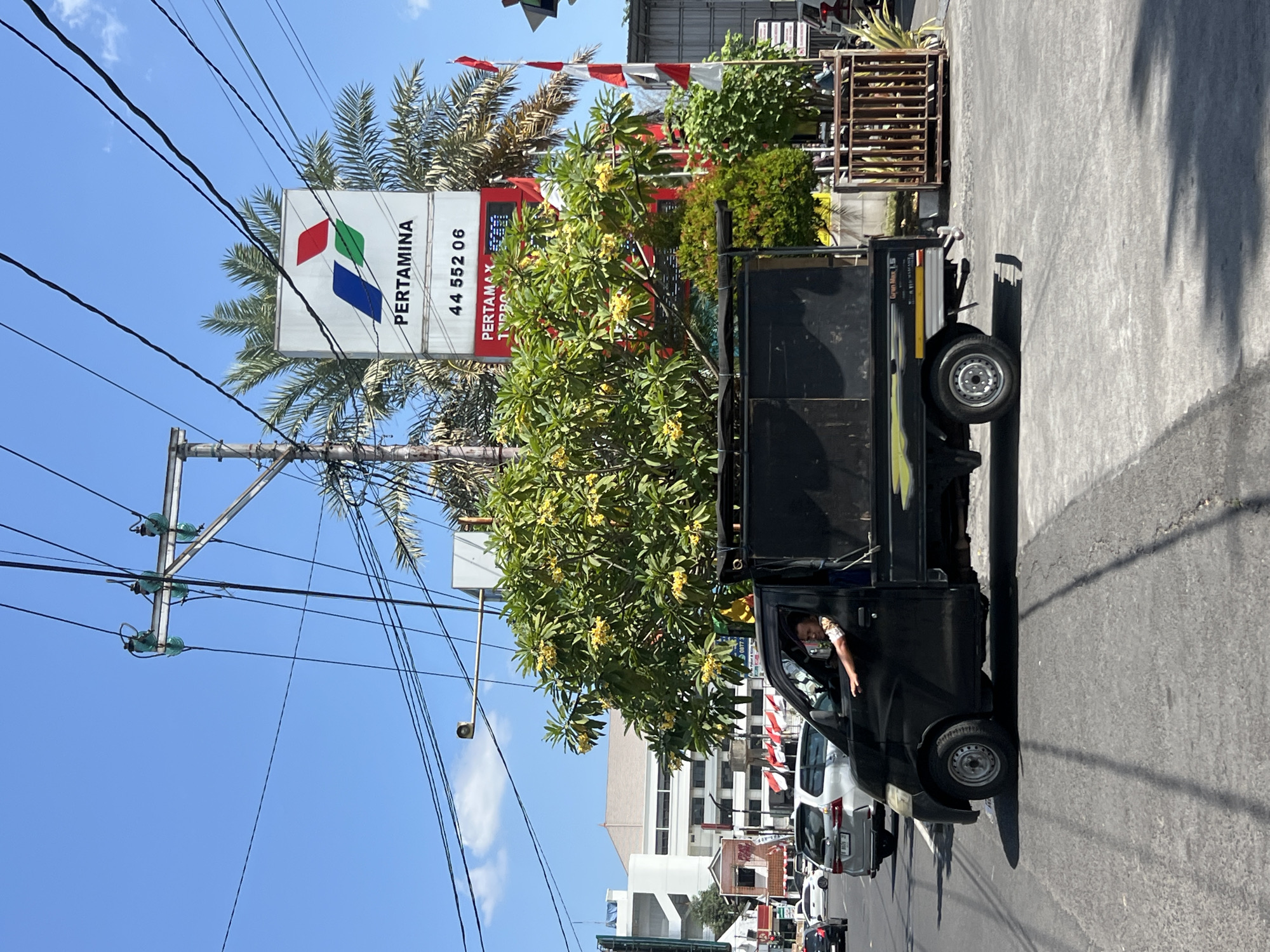 The image depicts a street scene in Yogyakarta, Indonesia, likely near a Pertamina gas station, given the prominent signage. A black pickup truck, with a man in the driver's seat, is turning at an intersection. The man is looking forward, with his right arm resting on the open driver's side window. The truck has a metal rack at the back. In the background, a Pertamina sign with the company logo and a phone number (44 552 06) is visible, along with palm trees and flowering trees. Several Indonesian flags (red and white) are planted along the roadside. Other vehicles, including a silver SUV and what appears to be another car, are present on the road, indicating moderate traffic. Buildings, including a white multi-story structure, are visible further in the background. The sky is clear and blue, suggesting a sunny day. Power lines and utility poles are a significant element in the foreground and midground.