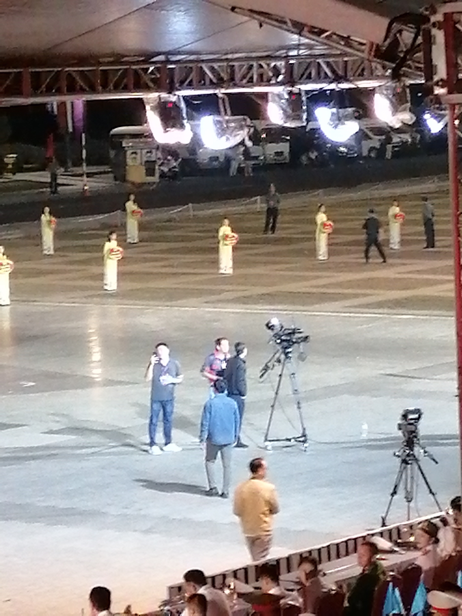 The media file shows an outdoor event with a large, flat paved area, likely a plaza or stage. A performance or ceremony is underway, featuring several individuals in traditional, light-colored robes holding red circular objects, possibly decorative or symbolic. They are arranged in a line across the paved area.
In the foreground, a man in a grey t-shirt and blue jeans is speaking into a microphone, possibly acting as a host or commentator. To his right, two other men are visible, one in a plaid shirt and another in a dark jacket. A camera on a tripod is set up nearby, suggesting a recording or broadcast is in progress. Further back, a person in a blue denim jacket and another in a yellow shirt are also present, with the latter facing away from the camera.
Above the paved area, a large, illuminated truss structure with spotlights is visible, indicating the event is taking place after dark or in a dimly lit environment. Several white vehicles, possibly vans or trucks, are parked behind the performers, some illuminated by the bright lights.
In the bottom portion of the image, an audience is seated in tiered seating. Some individuals are looking towards the event. The atmosphere appears to be formal and celebratory. While the exact time of day is not explicit, the presence of artificial lighting suggests it is evening or night. The location is identified as Da Lat, Vietnam, though no specific landmarks are clearly identifiable in the image. No visible text is present.