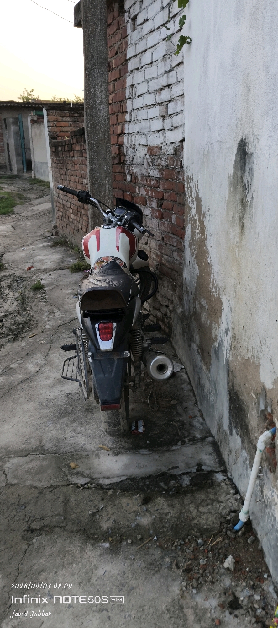 A motorcycle is parked in a narrow alleyway next to a weathered brick wall painted white in some sections and a rough concrete wall. The motorcycle, facing away from the camera, has a white and red fuel tank and a dark seat. Its exhaust pipe is visible on the right side. The ground is a mix of dirt and cracked concrete. In the background, a portion of a building with a brick wall and a blue door is visible. The image is timestamped 2026/09/08 08:09 with the text "Infinix NOTE 50S SG+" and "Javed Jabbar" overlaid. The location is identified as Bahera, India.