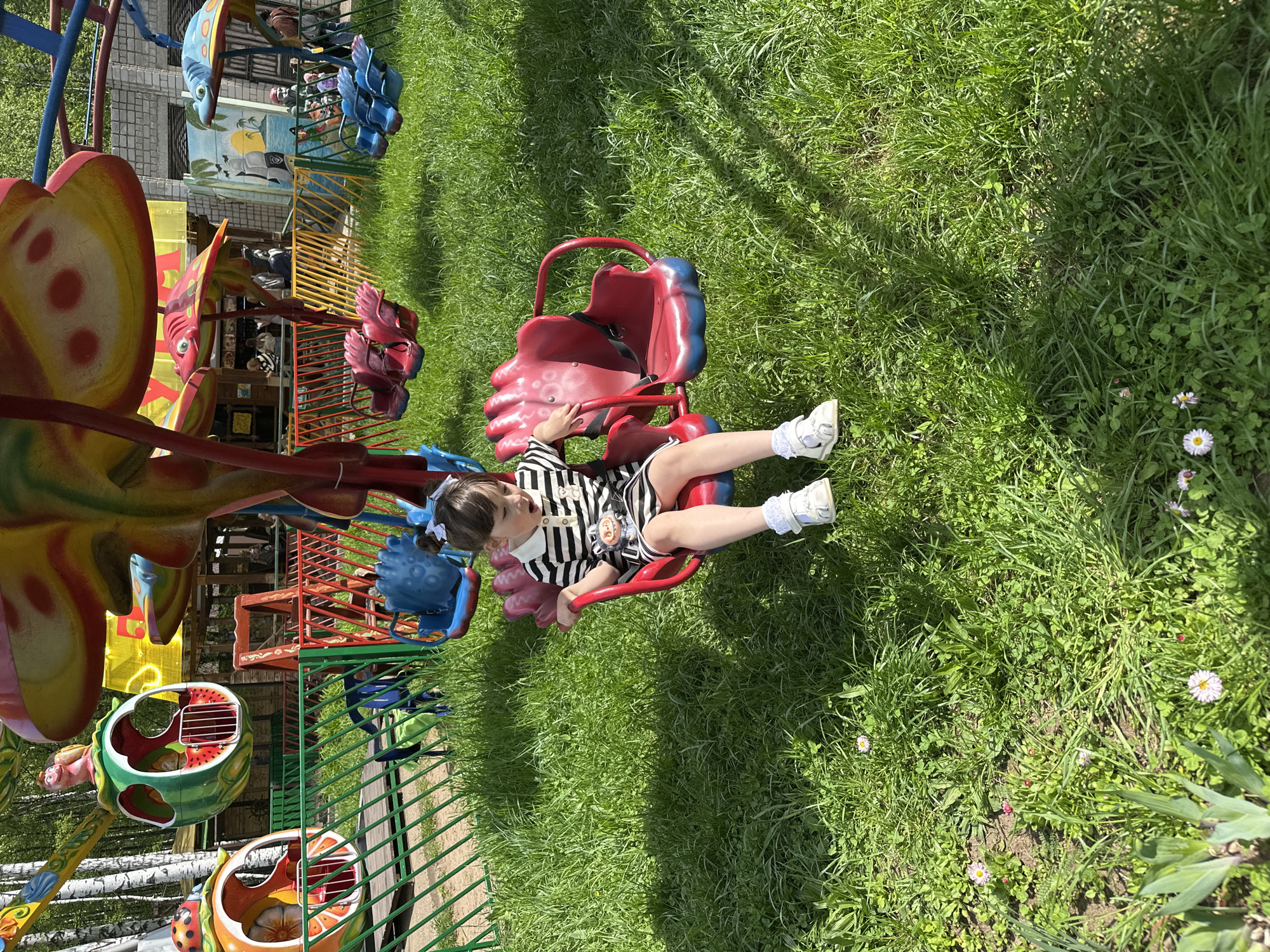 A young girl sits in a red and blue chair on a children's carousel at an amusement park. She wears a black and white striped outfit and white shoes. The carousel has other colorful, cartoon-like animal chairs. The scene is outdoors on a sunny day with green grass surrounding the carousel. This is Chaikovsky, Russia.