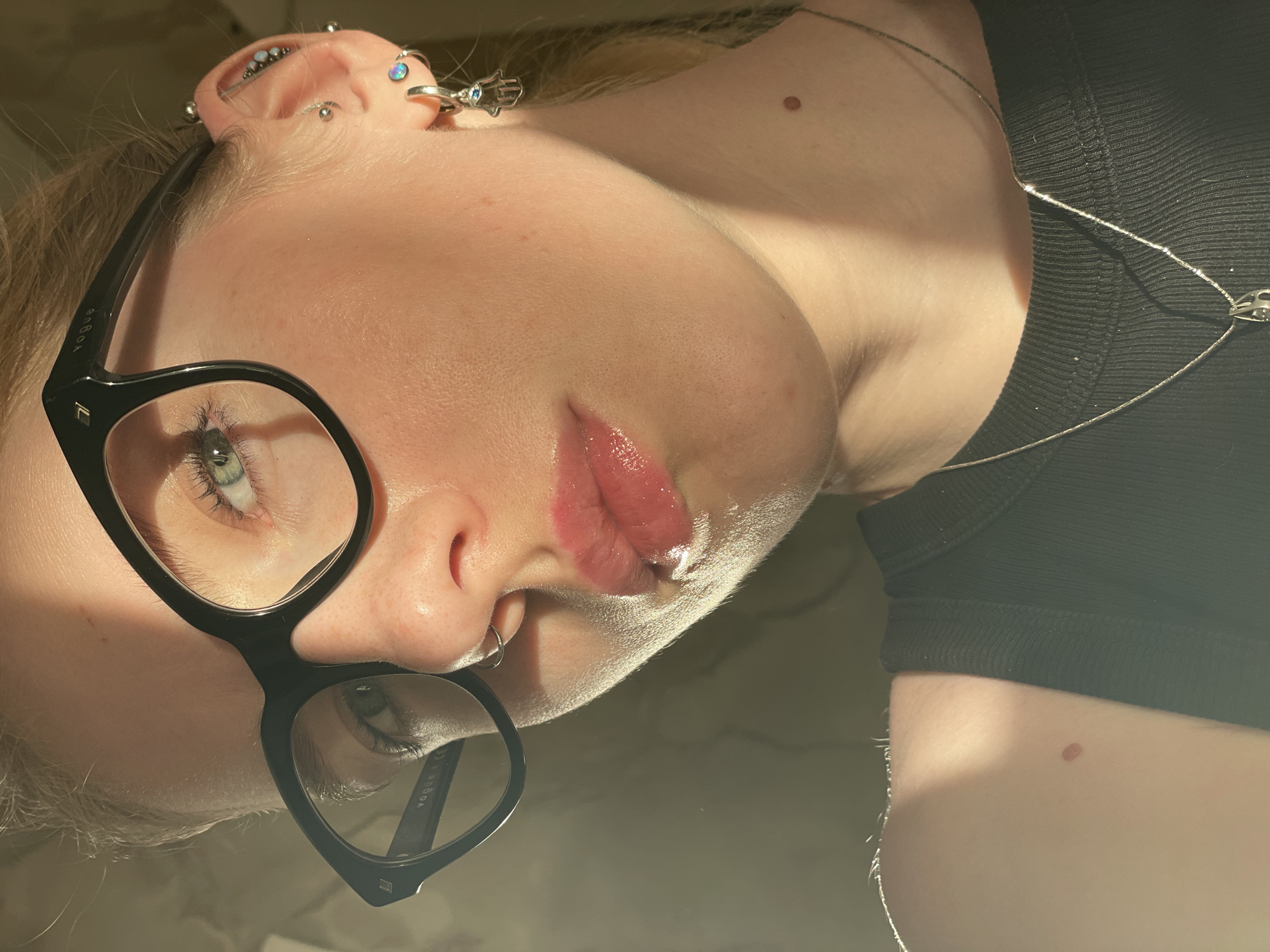 A close-up photograph captures a young person with fair skin, wearing black eyeglasses and a black top. The person is posed with their head tilted, presenting a view of their face, upper chest, and neck. The person has multiple piercings on their left ear. A silver chain necklace is visible around the person's neck. The lips appear glossy. The scene is illuminated by a natural light source, creating highlights and shadows on the skin. There is no indication of the city of Izhevsk, Russia, in the image.