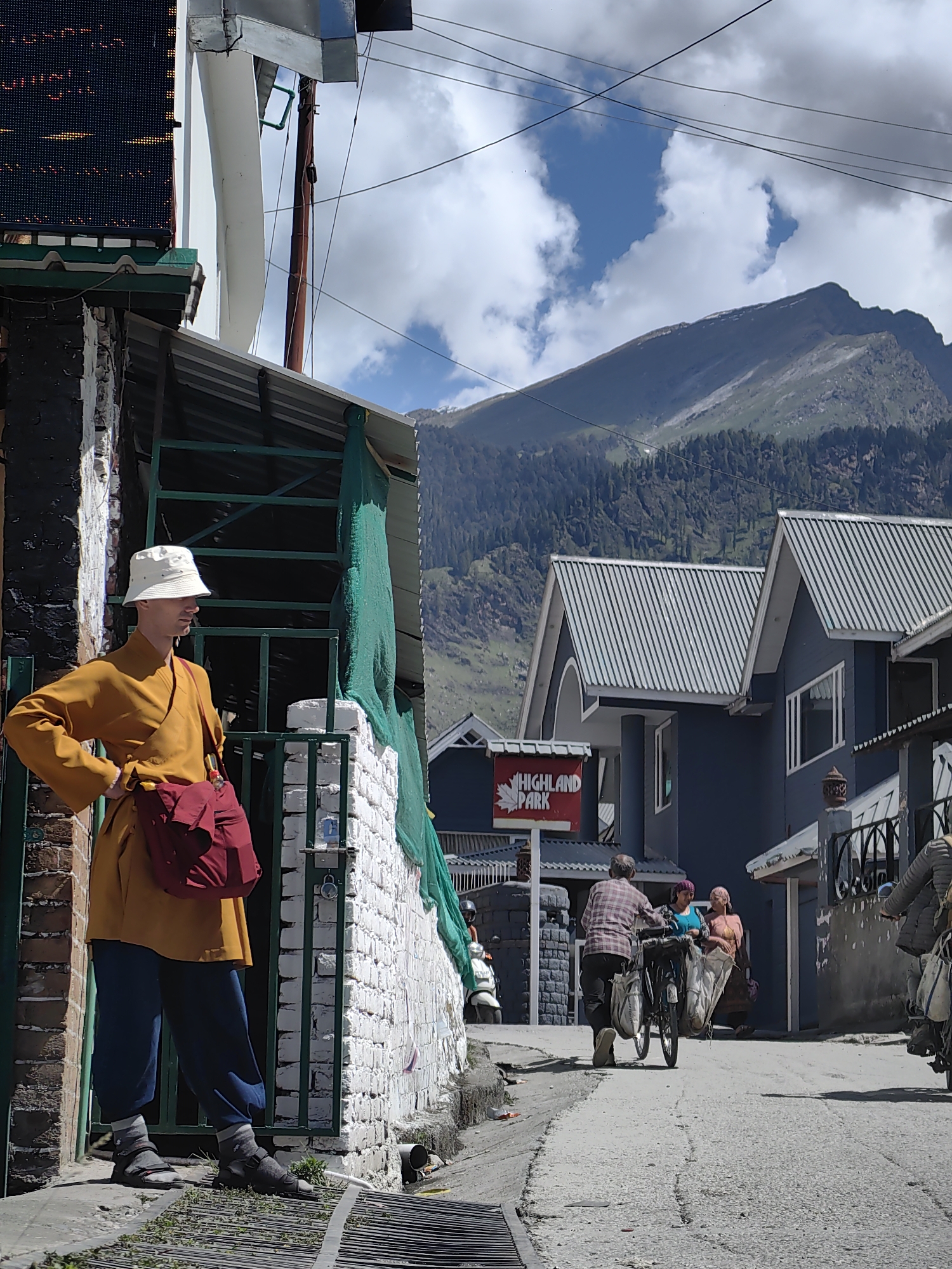 The image depicts a street scene in Vashisht, India, with a prominent mountain range in the background. In the foreground, a person dressed in a saffron robe and a white bucket hat stands with their hand on their hip, carrying a red bag. To the right, a street runs uphill, with a man pushing a bicycle laden with two large bags. Two women stand nearby, one in a blue top and the other in a pink top. Further up the street, several dark blue buildings with silver corrugated roofs are visible, one bearing a red sign with white text that reads "HIGHLAND PARK." The sky is partially cloudy, with patches of blue. The environment appears to be a rural or semi-rural area, likely a tourist destination given the "Highland Park" sign and the presence of visitors. The overall atmosphere is one of everyday life in a mountainous region.