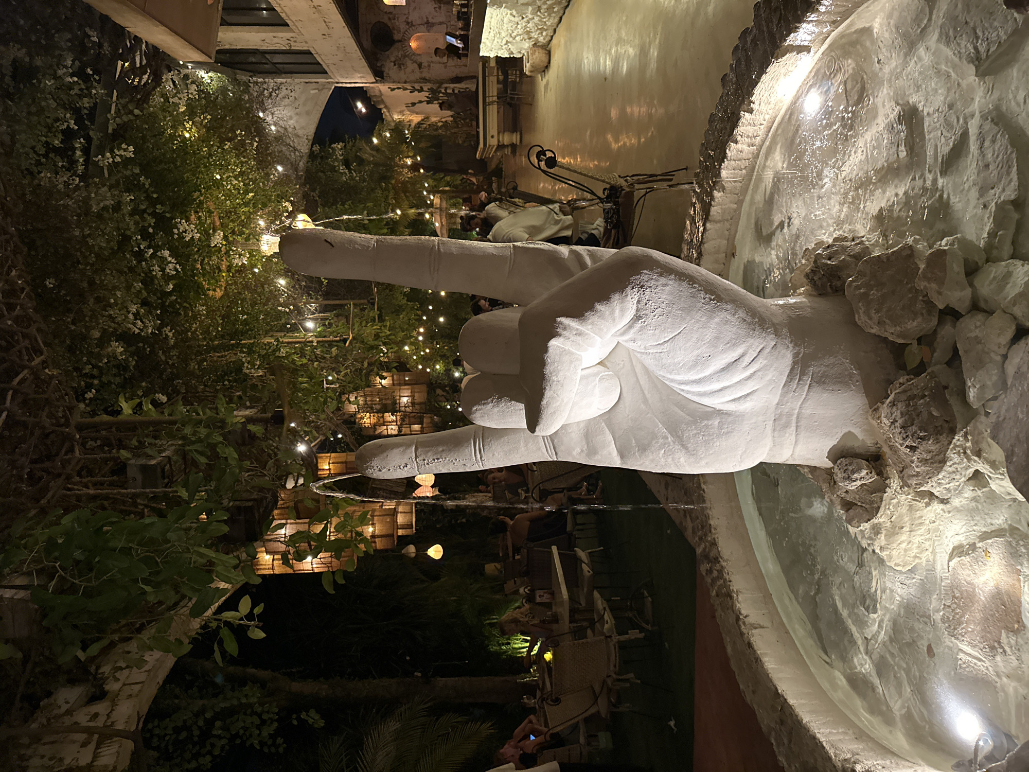 This image depicts an evening scene at an outdoor or semi-outdoor establishment, likely a restaurant, bar, or courtyard. In the foreground, a prominent white sculpture of a hand making the "rock on" or "devil horns" gesture serves as a fountain, with water cascading from its fingertips into a light-colored, rocky basin. The fountain and its basin are illuminated, highlighting the texture of the stone and the movement of the water.
The background is rich with greenery, featuring lush trees and shrubs, some adorned with small white flowers. Numerous string lights and decorative lanterns are strung throughout the foliage and structures, casting a warm, inviting glow over the entire area. Several people are visible sitting at tables and chairs, scattered among the greenery, suggesting they are patrons socializing or dining. Architectural elements in the background, including light-colored stone walls and arches, hint at a Mediterranean or similar architectural style. The time of day is clearly night, with the dark sky contrasting with the artificial lighting. The overall atmosphere appears relaxed and festive. No visible text is present in the image.