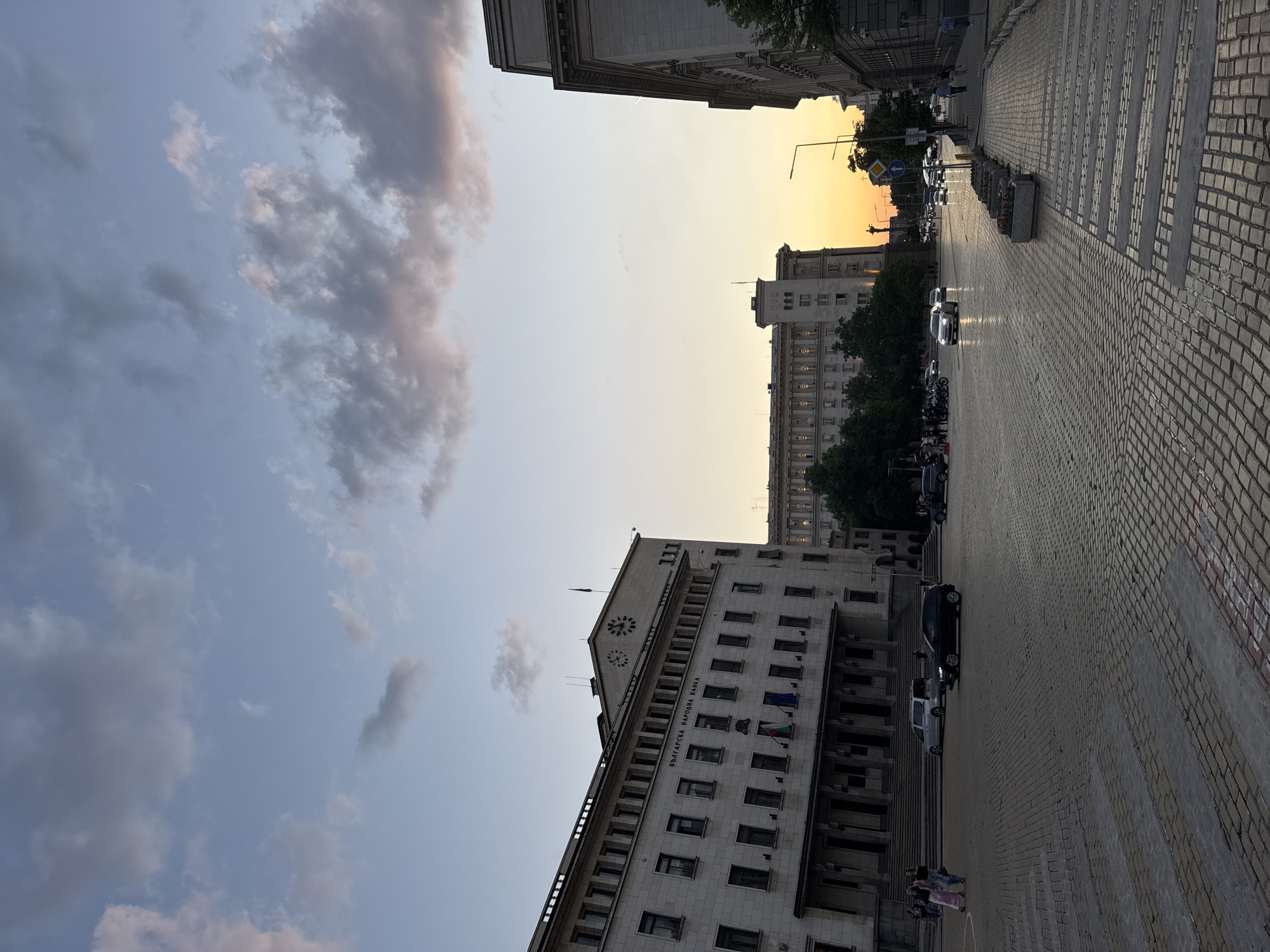 The image captures a street scene in Sofia, Bulgaria. The Bulgarian National Bank building is visible, situated on a cobblestone street. The sky transitions from dusk to evening, with clouds present. Several vehicles are parked along the street. Pedestrians are present on the sidewalk. Buildings line the street, and a street light is visible in the distance.