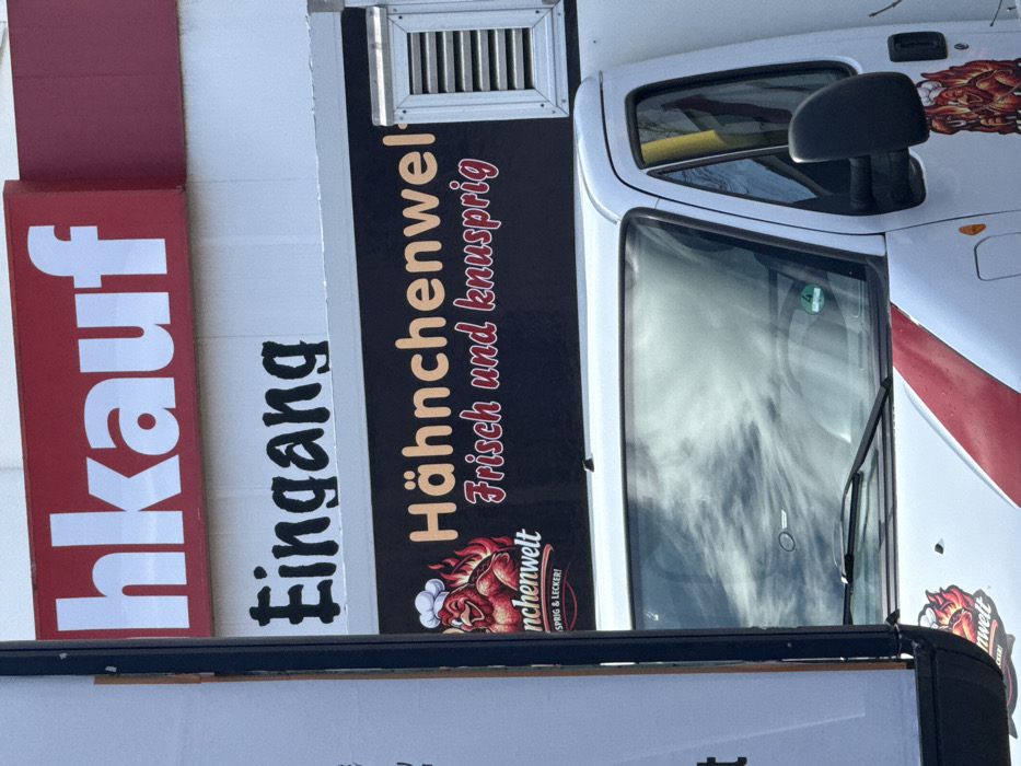 The image depicts an outdoor commercial scene at the Kaufland supermarket on Holländische Straße 135, 34246 Vellmar, Germany.
In the upper portion, a red sign with white capitalized letters, partially visible, displays "...hkauf," indicating a retail establishment. Directly beneath it is a white sign with black stylized text that reads "Eingang" (German for Entrance).
Dominating the lower and central foreground is the front half of a white food truck. The truck's windshield reflects a cloudy sky. A green environmental sticker is affixed to the lower left side of the windshield. A black sign on the side of the truck, positioned above the cab, features orange capitalized text "Hähnchenwelt" (Chicken World). Below this, in red script, are the words "Frisch und knusprig" (Fresh and crispy). To the left of this text, a cartoon logo depicts a red, flame-outlined chicken wearing a white chef's hat. Further down the truck's side, partially obscured by a black vertical post on the left, additional text is visible, including "nchenwelt" and "SPRIG & LECKER!" (Crispy & Delicious!). A silver louvered vent is visible on the upper right side of the truck's black sign. No individuals or specific actions are visible within the frame.