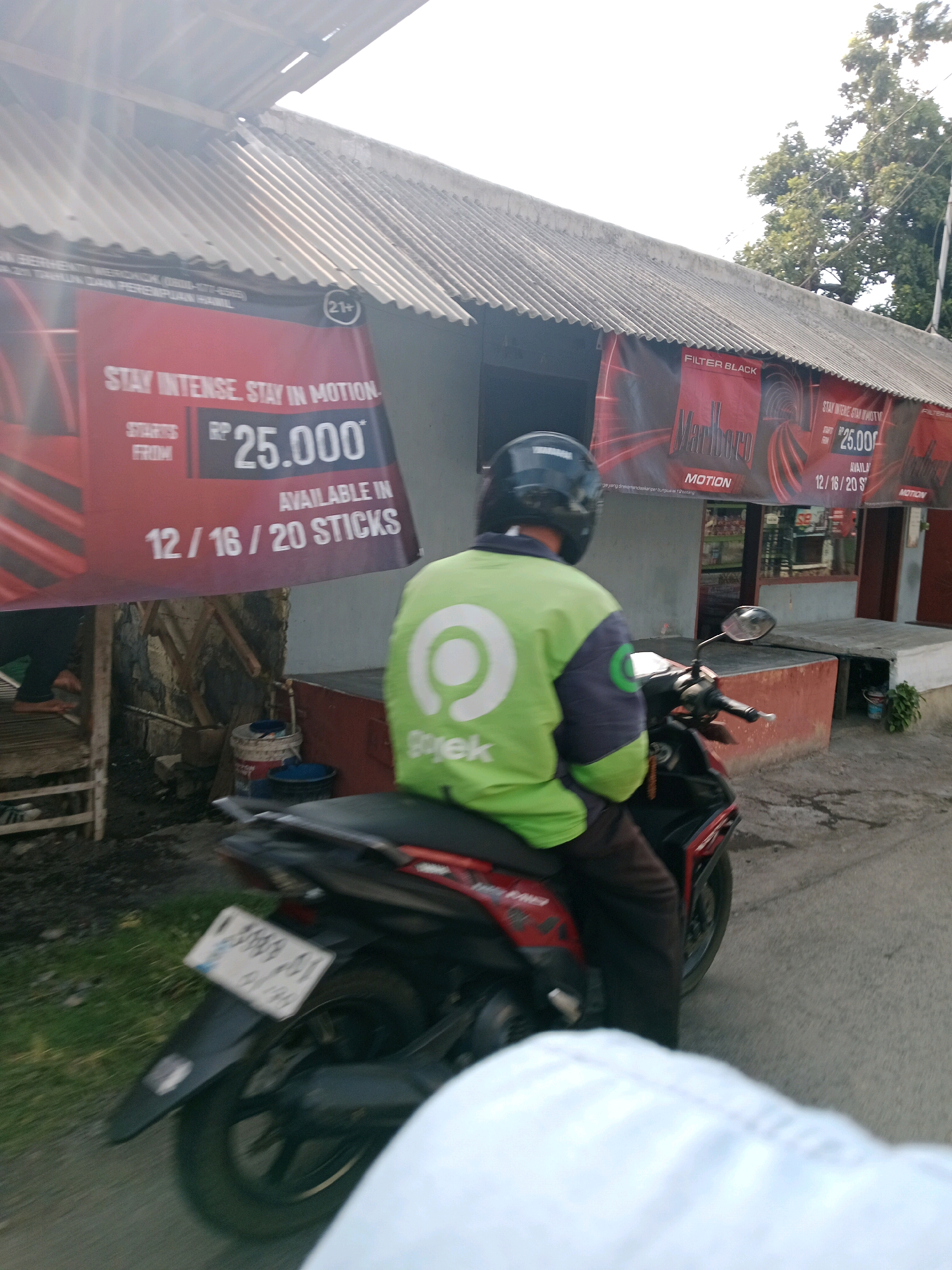 The image depicts a street scene in Probolinggo, Indonesia. A person wearing a green and dark blue jacket with a helmet is riding a motorcycle. The jacket prominently features a white circular logo with a smaller white circle inside and the word "Gojek" written below it, indicating they are a Gojek driver. The motorcycle is black and red, with a visible license plate that reads "L 3668 AY". In the background, there is a building with a corrugated metal roof, and several red banners advertising "Marlboro" cigarettes. One banner also shows "STAY INTENSE. STAY IN MOTION." and prices like "Rp 25.000". The environment appears to be outdoors, with some vegetation and a light-colored sky. The image seems to be taken from another vehicle, as a white blurred surface is visible in the foreground.