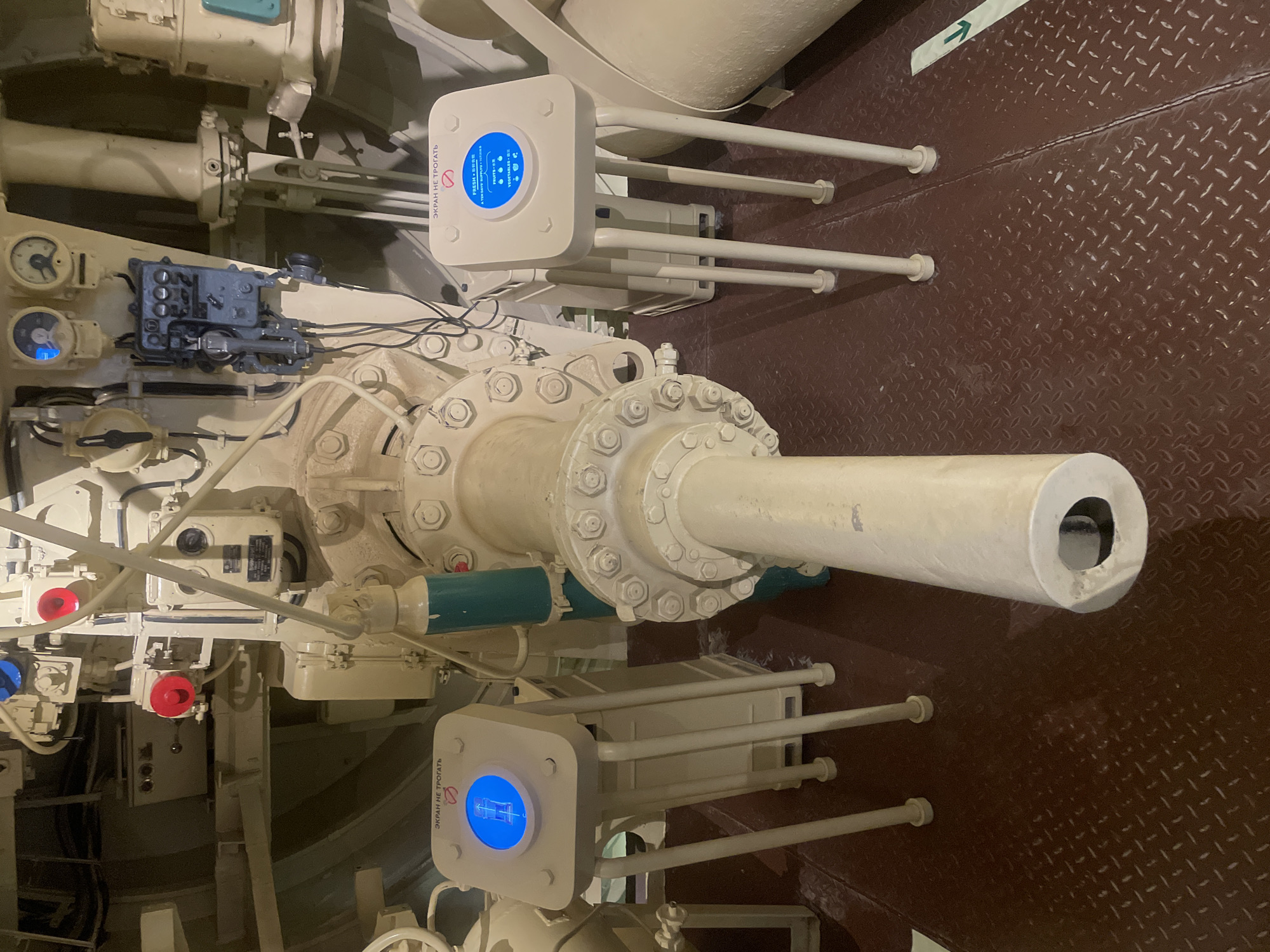 The image shows the interior of a vessel, possibly a submarine, with complex machinery and control panels. In the foreground, a large cylindrical object, likely part of a gun or torpedo tube, extends from the center towards the viewer. Two white, square interactive display units with blue screens are positioned on either side of this central object. The screens display text in Russian, "ЭКРАН НЕ ТРОГАТЬ" (Do not touch the screen), and graphics related to fresh produce. The environment appears industrial, with exposed pipes, gauges, and electrical components painted in off-white and beige tones. The floor is a dark red, diamond-patterned metal plating. A green directional arrow is visible on the floor to the right of the frame. The overall impression is of a well-maintained, but functional, engineering space. This scene is located within the Kronstadt Museum-Preserve, an open-air museum complex in Kronstadt, Russia, which preserves naval history.