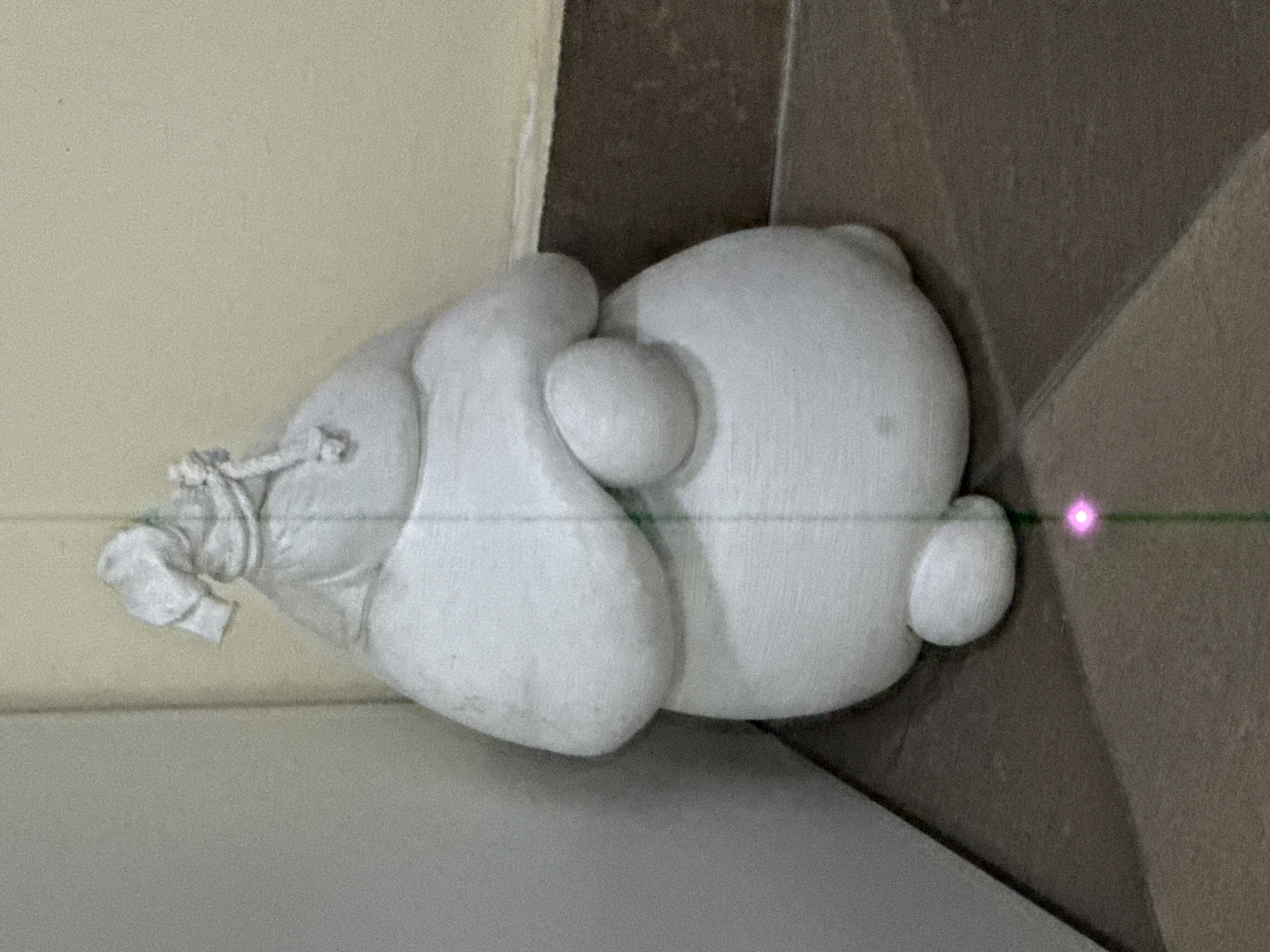 A white, plump sculpture resembling a character is positioned on a surface, possibly a floor, with the edge of a wall visible. The sculpture has a rounded head-like form and what appears to be a drawstring bag attached to the side. A green laser line with a pink dot crosses the image, seemingly aimed at the sculpture. The surface beneath the sculpture is a two-tone brown, suggesting tile or a similar material. No information about Khimki, Russia, can be found in the image.