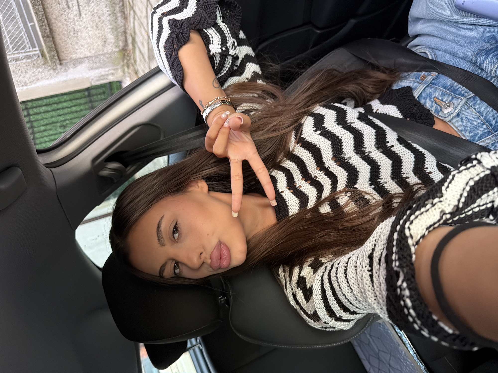 A young woman with long, dark brown hair is in the driver's seat of a car, taking a selfie. She is wearing a black and white striped sweater and light blue denim jeans. Her left hand is held up, forming a peace sign, and she is wearing several bracelets on her left wrist. Her lips are slightly parted, and her eyes are looking at the camera. The interior of the car is visible, including the black leather seats and seatbelt. Outside the car window, a grey building with a metal grate on a window and some greenery can be seen, consistent with a street view in Karnobat, Bulgaria.