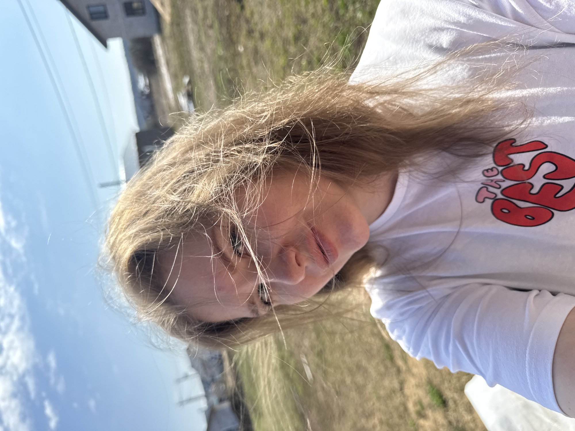 The image presents a close-up, chest-up portrait of a young person, likely female, situated outdoors on a bright, sunny day. The individual has long, light brown or blonde hair, appearing somewhat disheveled or wind-blown, with sunlight catching and highlighting individual strands. She is wearing a white long-sleeved t-shirt featuring a prominent red graphic design on the front. The design clearly spells out "THE BEST" in a stylized, bubbly font, with "THE" positioned above "BEST."
The person's face is angled slightly, revealing a relaxed expression with a subtle, gentle smile. Strong sunlight illuminates her, creating highlights on her hair and skin.
The background is somewhat blurred but depicts an open outdoor setting. Above, a clear blue sky with a few wispy clouds is visible. To the left, a portion of a building, possibly a house or other structure, can be faintly seen. The ground behind the person appears to be covered in sparse, dry grass or dirt, suggesting a rural or suburban environment. The robust sunlight indicates that the photo was taken during the daytime, likely midday or early afternoon. The overall mood conveyed is one of casual comfort and pleasant weather. The location context for this media file is Gornyi (Dvurechenskoe S/P), Russia.