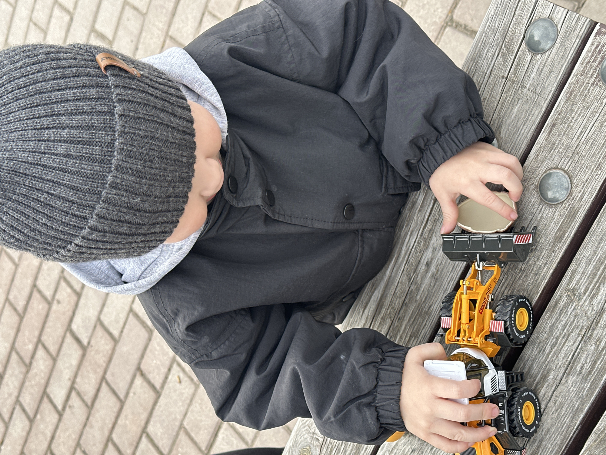 A child wearing a gray knit hat, a gray hoodie, and a black jacket is sitting on a wooden bench. The child is holding and playing with a yellow and black toy loader. The bench is on a brick-paved surface. The location is not discernible as Votkinsk, Russia, based solely on the provided image.
