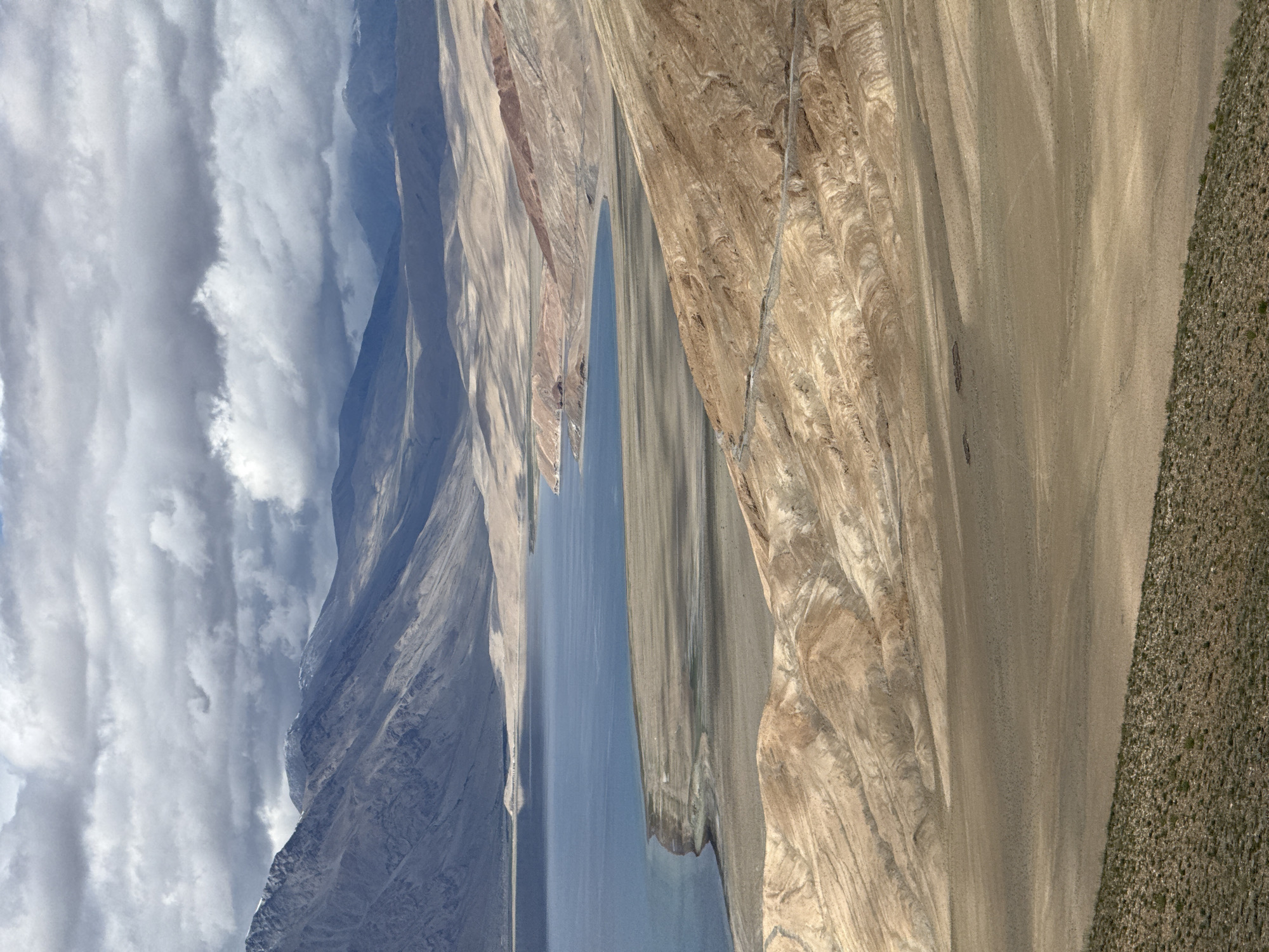 The image displays a panoramic view of a vast, arid landscape surrounding a large, blue body of water. The water's edge is defined by a sandy, somewhat barren shoreline, transitioning into rolling, tan-colored hills and mountains. These geological formations exhibit layers and striations, suggesting erosion and time. A winding road is visible snaking its way up one of the hillsides, indicating human presence in this remote environment. In the background, taller, majestic mountains rise, their peaks partially obscured by dramatic, grey and white clouds that fill the sky. The overall impression is one of rugged, natural beauty and solitude. The scene is consistent with high-altitude desert or steppe regions.