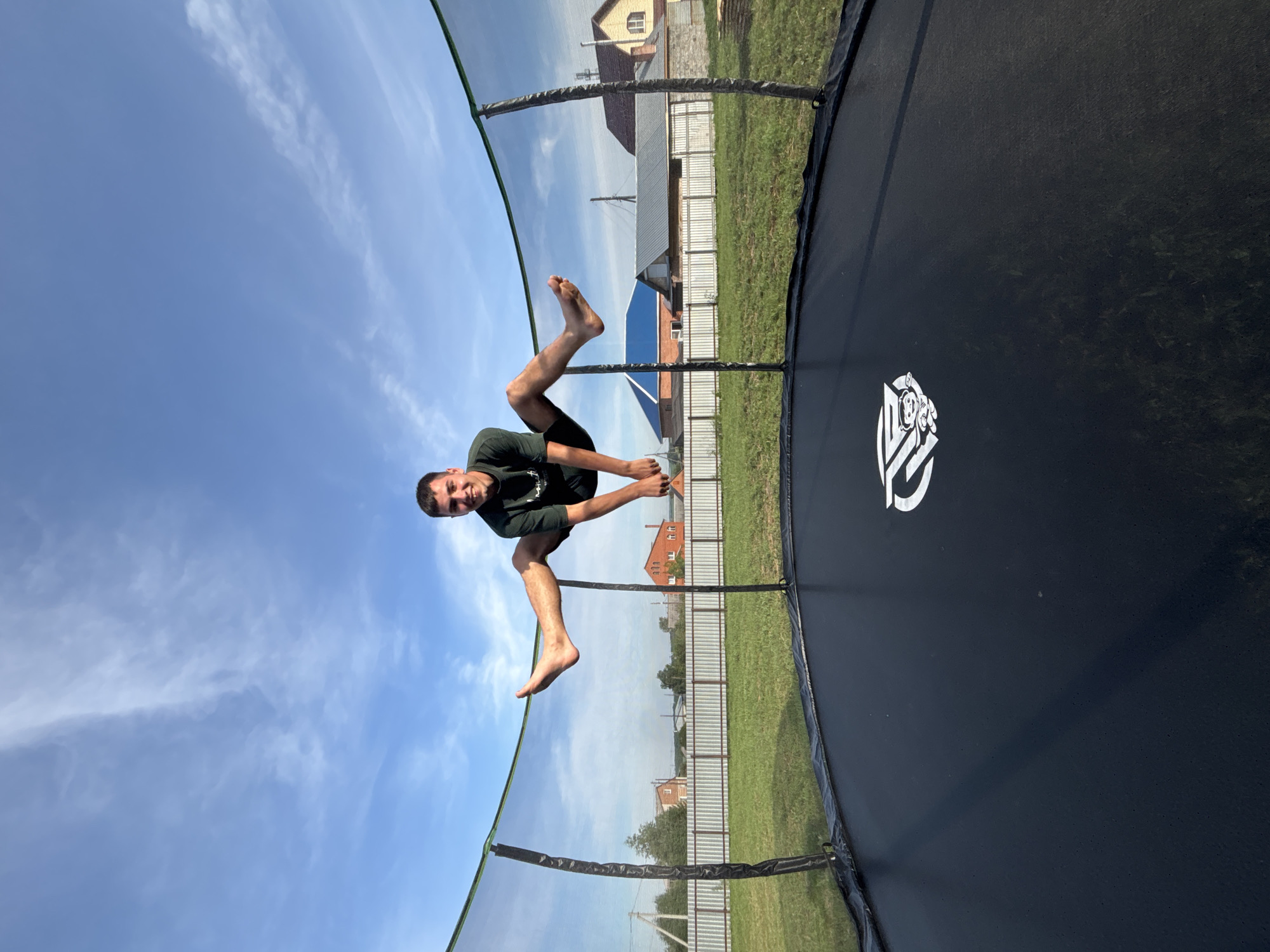 In the city of Vishnevka, Russia, a young man with short, dark hair and a light complexion is captured mid-air, performing a split jump on a black trampoline. He is wearing a dark green t-shirt and dark shorts. The trampoline is enclosed by a green safety net, with black poles supporting it. The background reveals a rural setting with a green grassy field, a white fence, and several houses with red and blue roofs. The sky above is a bright blue with scattered white clouds, suggesting it is daytime.