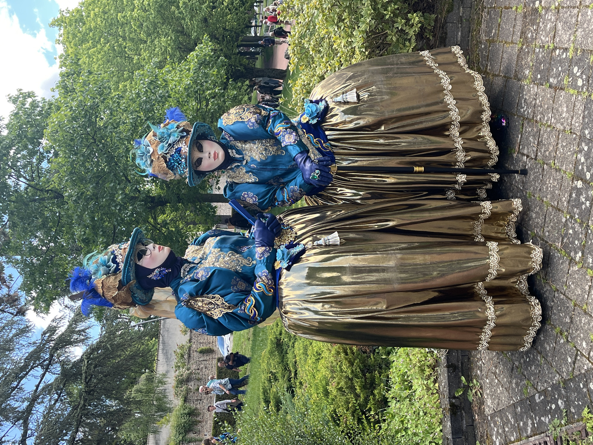 Two individuals in elaborate traditional carnival costumes stand side-by-side on a cobblestone path. The costumes consist of gold, floor-length skirts with ornate lace trim, complemented by deep blue jackets adorned with gold embroidery, matching gloves, and hats decorated with feathers and beads. Both wear masks, obscuring their faces. One holds a black cane.
The scene is set in a park or garden environment, with green trees and bushes in the background. Other individuals are visible in the background, suggesting a public event or gathering. The cobblestone path is grey and worn. Based on available information, the location is Saint-Galmier, France.