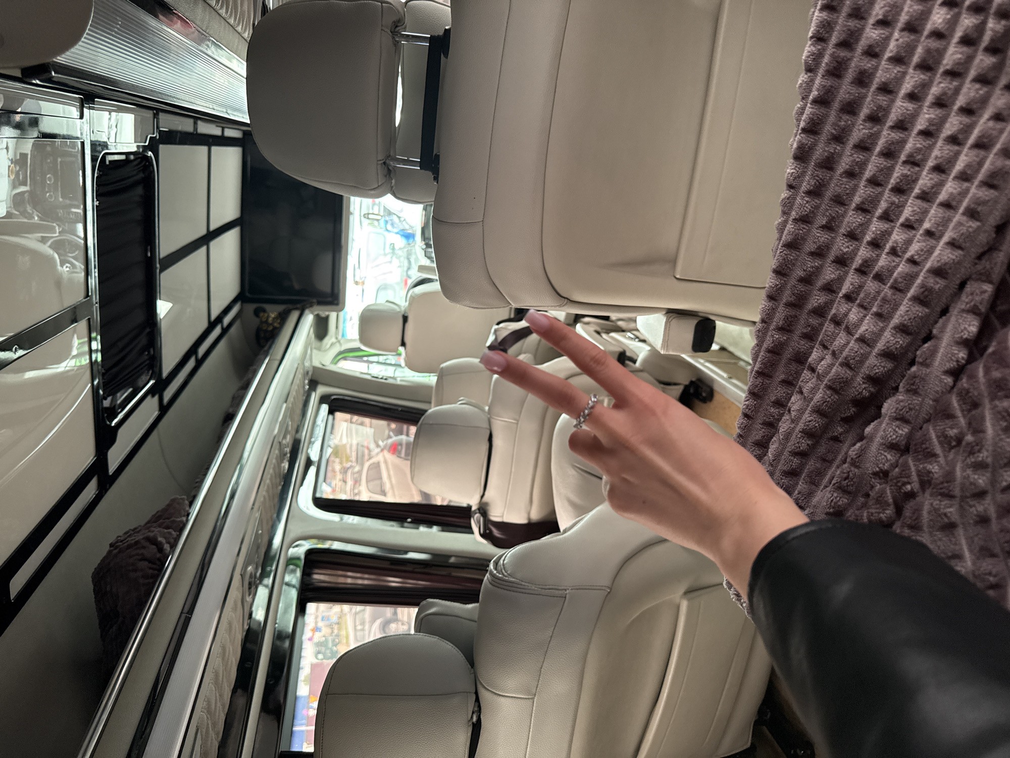 The image shows the interior of a vehicle, possibly a van or bus, with multiple rows of white leather seats. A person's hand is visible in the foreground, making a peace sign. The vehicle appears to be in Bielany Wrocławskie, Poland, as suggested by the view through the windows showing urban or suburban landscape with buildings and other vehicles, though specific landmarks confirming this location are not discernible. A dark, textured fabric is draped nearby. A television or display is mounted on the wall opposite the seats.