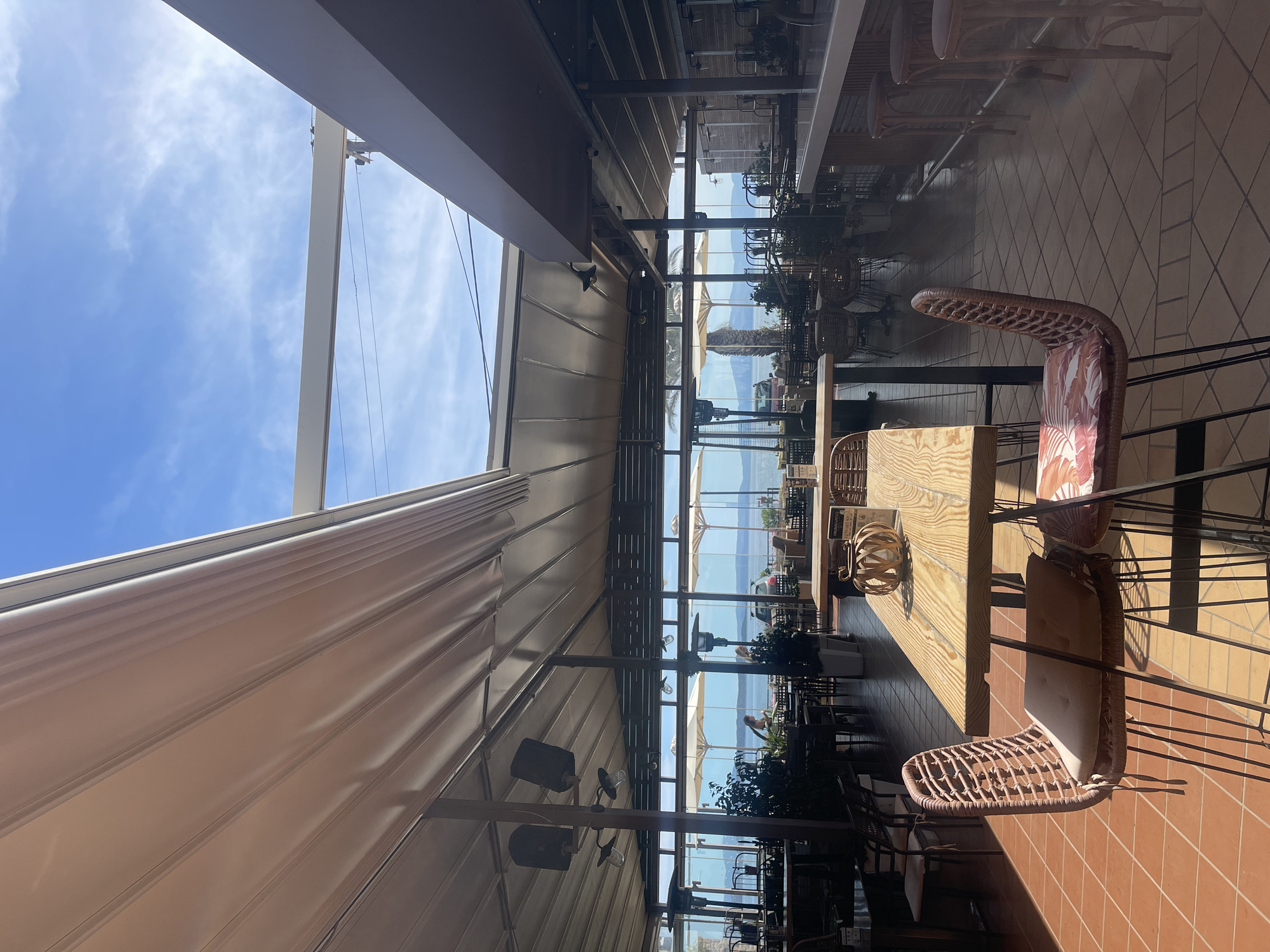 The image depicts the interior of a restaurant in Nea Makri Municipal Unit, Greece. The scene is viewed from the perspective of a table. Two chairs are visible at the table. Beyond the table, there is a glass wall that looks out over the Aegean Sea. Some people and vegetation can be seen along the coast. Above the interior space, there is a retractable awning. The sky is visible through the opening above the awning.