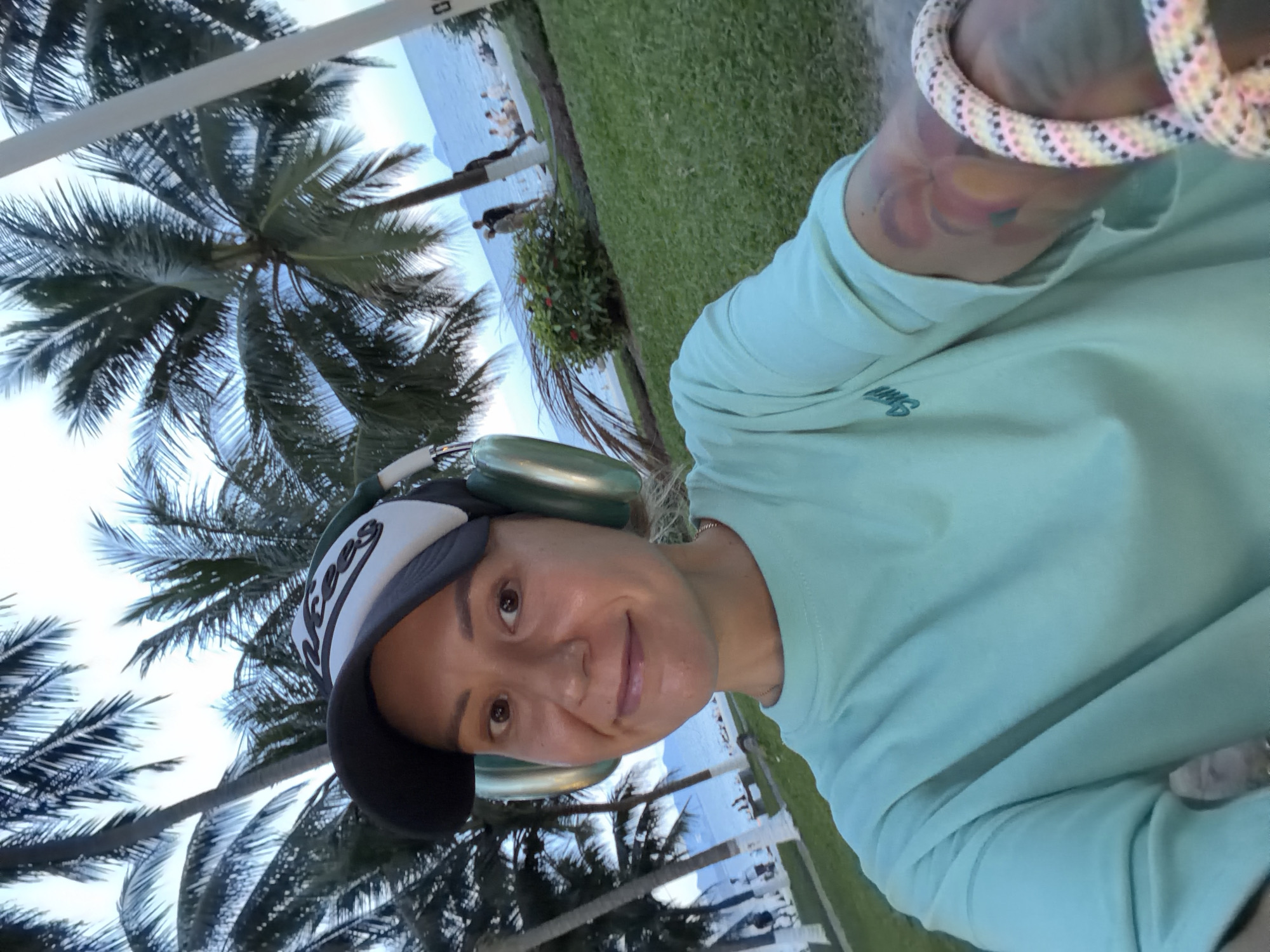 A woman is taking a selfie in an outdoor setting. She is wearing a light blue t-shirt, a baseball cap with a white and black logo, and large, light green headphones. She has tattoos on her left arm, visible on her forearm and upper arm. She is smiling at the camera.
Behind her, palm trees are prominent, with their fronds reaching towards a cloudy sky. The background shows a grassy area leading down to a beach and the ocean. There are several people in the distance on the beach and near the water, some appearing to be sitting or walking. The overall environment suggests a tropical or coastal location. The image does not provide enough information to definitively place the location in Nha Trang Ward, Vietnam.