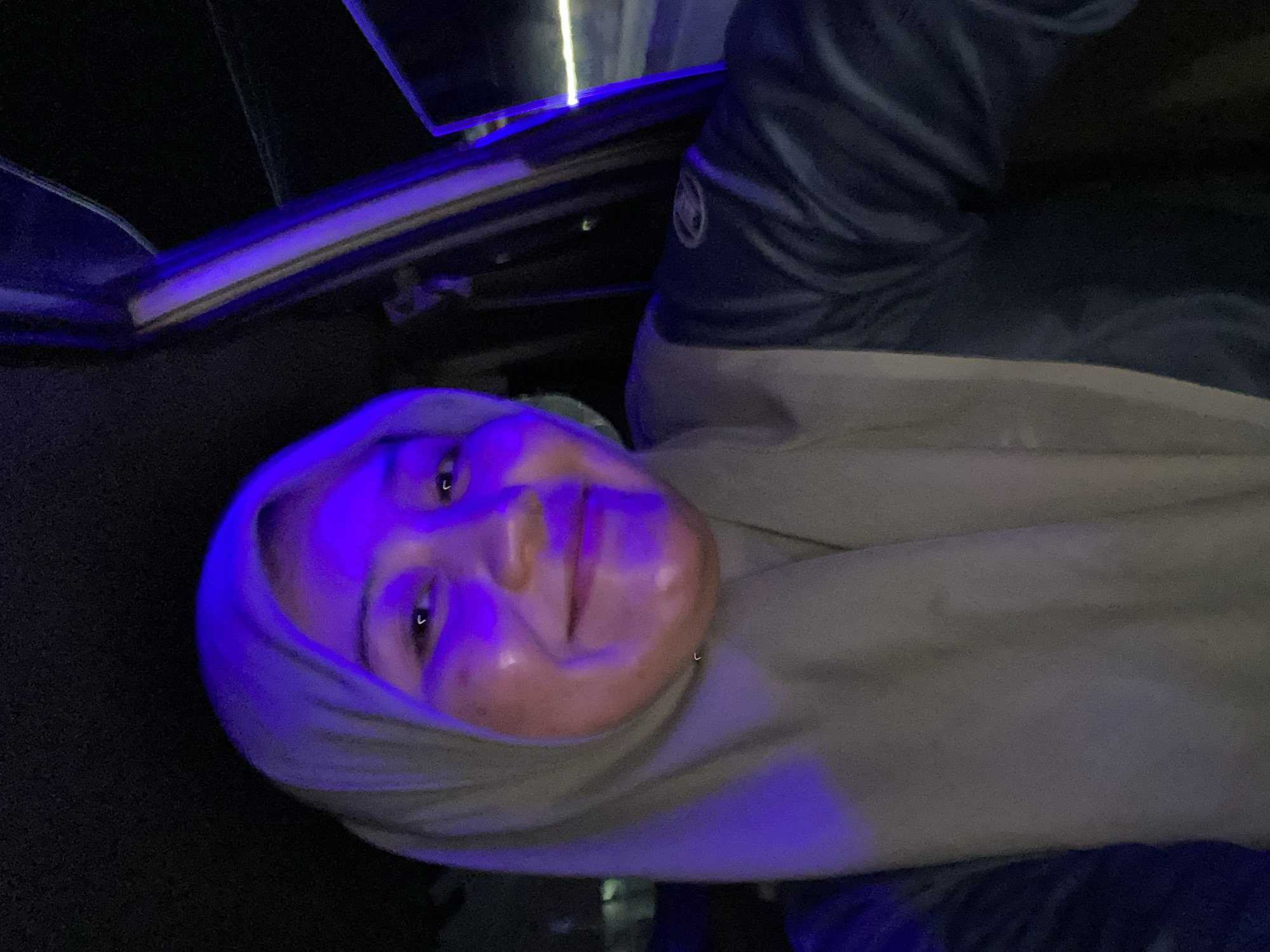 A woman wearing a beige hijab and a dark blue jacket is captured in a selfie from within a vehicle. The lighting is predominantly blue, casting a distinct glow on her face, highlighting her smile. The background is dark, suggesting it is nighttime, with some blurry lights visible through the car window on the right. The location is identified as Sungai Paring, Indonesia.