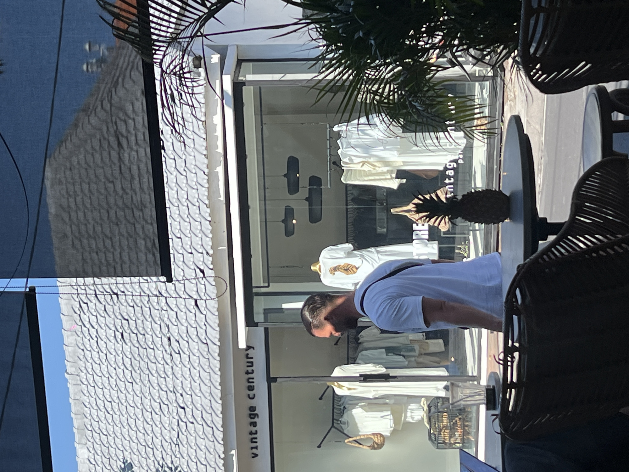 The image shows a street view in North Kuta, Indonesia. The "vintage century" store, displaying white clothing, is visible through the shop window. A man in a white t-shirt is reflected in the window, his back turned toward the viewer. A black rattan chair and table, bearing a pineapple decoration, are prominent in the foreground, partially obstructing the view of the store. The building has a scale patterned roof, and a palm tree hangs in the top of the frame.