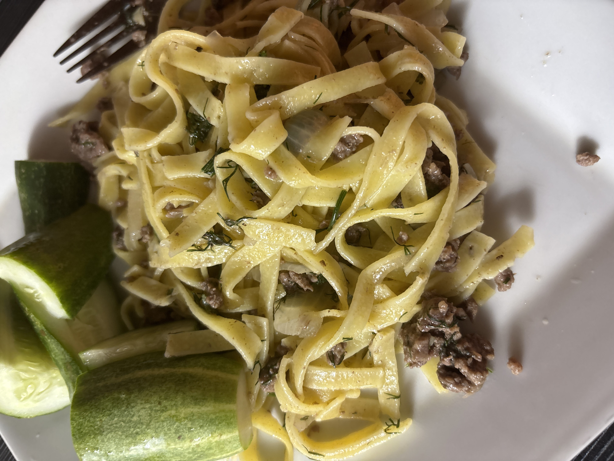 A top-down shot shows a white plate containing a serving of tagliatelle pasta mixed with ground meat and dill. To the left, there are three thick slices of cucumber. A metal fork is visible at the top left corner. The photograph appears to be taken indoors, likely in a home or restaurant in บ้านบางรักษ์, Thailand.