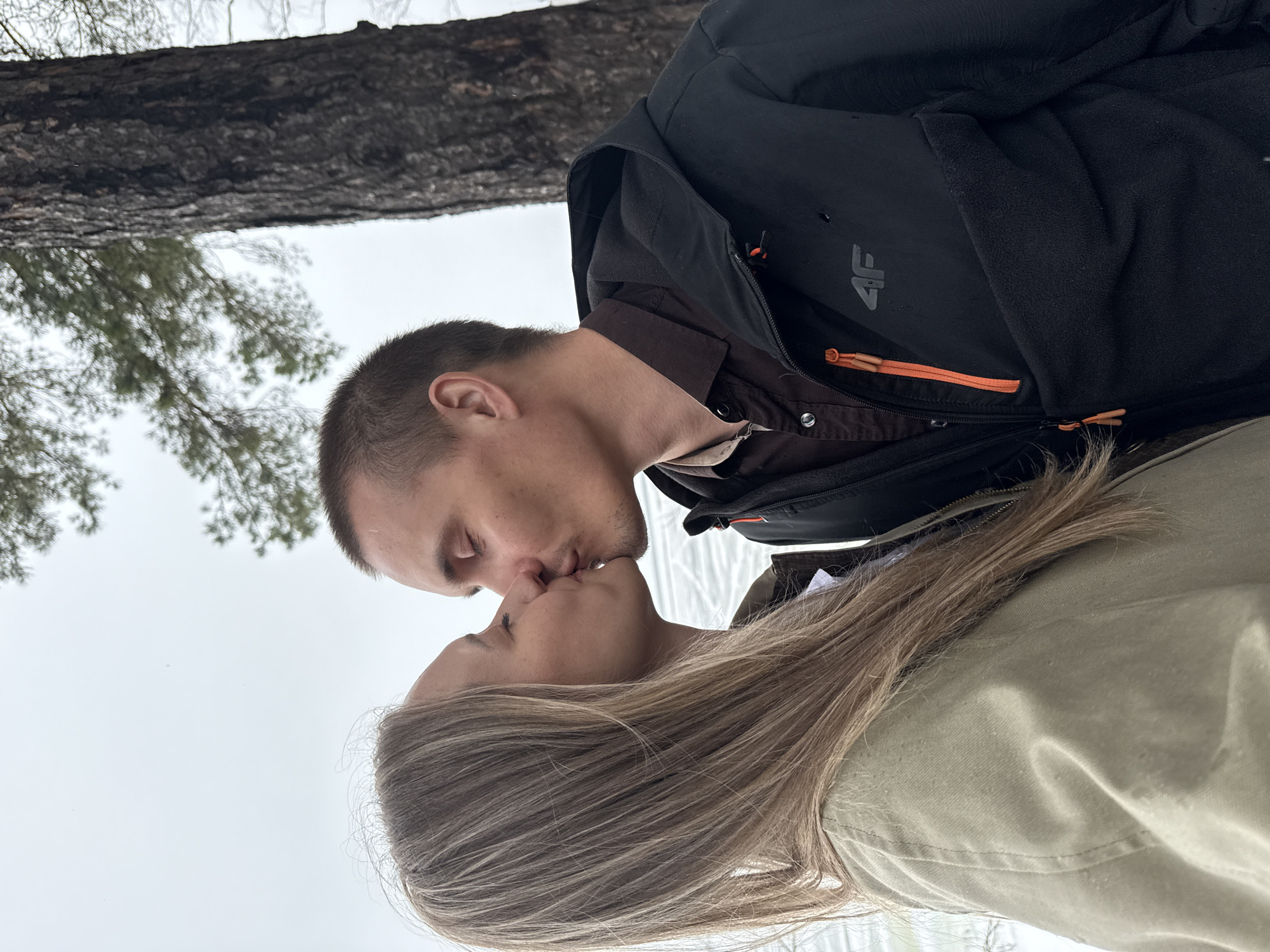 A man and a woman are kissing. The man is wearing a black jacket with an orange zipper and a brown shirt. The woman has long, light brown hair and is wearing a green jacket. There is a tree and some sky visible in the background. The location is Chaikovsky, Russia.