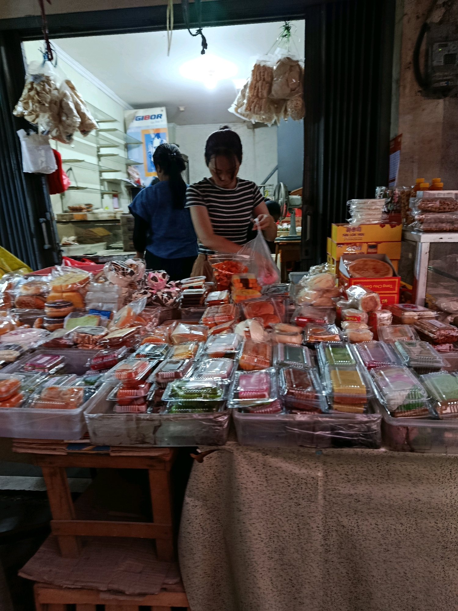 In Bekasi, Indonesia, a street vendor displays a variety of packaged food items on a table. The table is covered with a textured cloth and is supported by a wooden stand. Numerous plastic containers filled with colorful sweets and snacks are arranged on the table. Behind the table, two women are visible. One woman, wearing a striped shirt, is in the process of handling a plastic bag of food. The other woman, in a blue shirt, is partially obscured. The background shows the interior of a shop with shelves and a sign that reads "GIBOR." The scene appears to be an outdoor market or roadside stall.