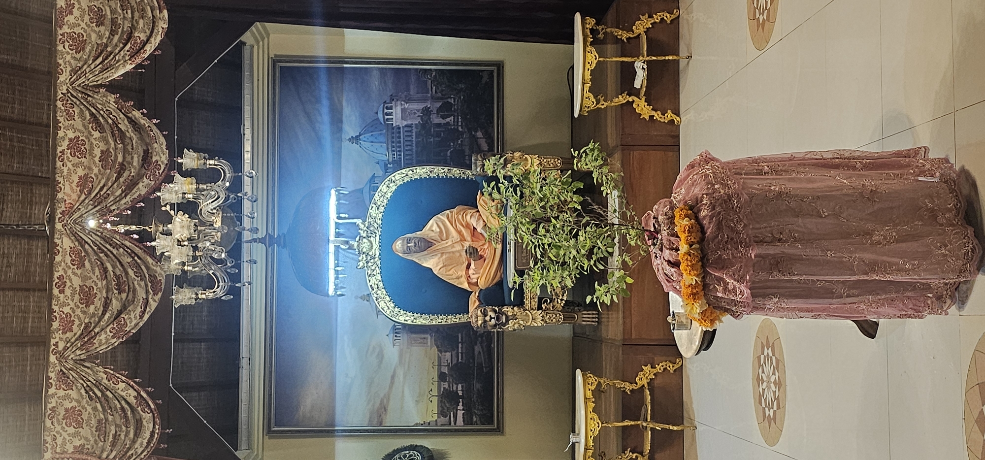 The image shows an interior view of a room in Denpasar, Indonesia. In the background is a framed image depicting a man in an orange robe seated on a blue chair against a backdrop of a building with a dome. In front of the framed image is a potted plant. To the left is a chandelier hanging from the ceiling, adjacent to decorative curtains. To the right is a draped table with a garland around it, set on a tiled floor featuring oval designs.