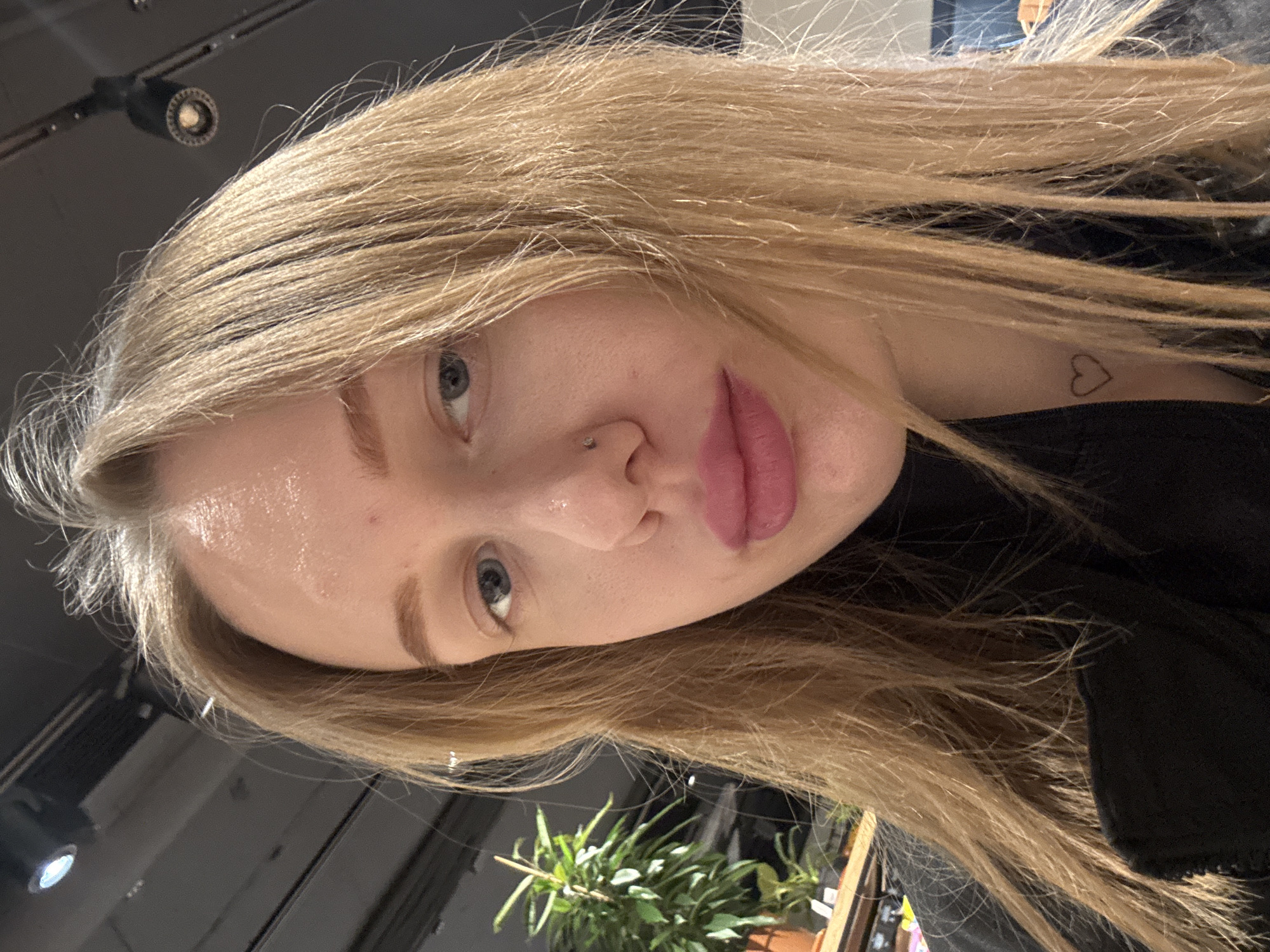 A close-up, angled selfie of a fair-skinned woman with light blonde hair and blue eyes. She has a piercing on the left side of her nose. A small heart tattoo is visible on her chest, near her neck. She is wearing a black garment. In the background, a potted green plant and a dark ceiling with track lighting fixtures are visible. The location in Rostov-on-Don, Russia, cannot be determined from the image.