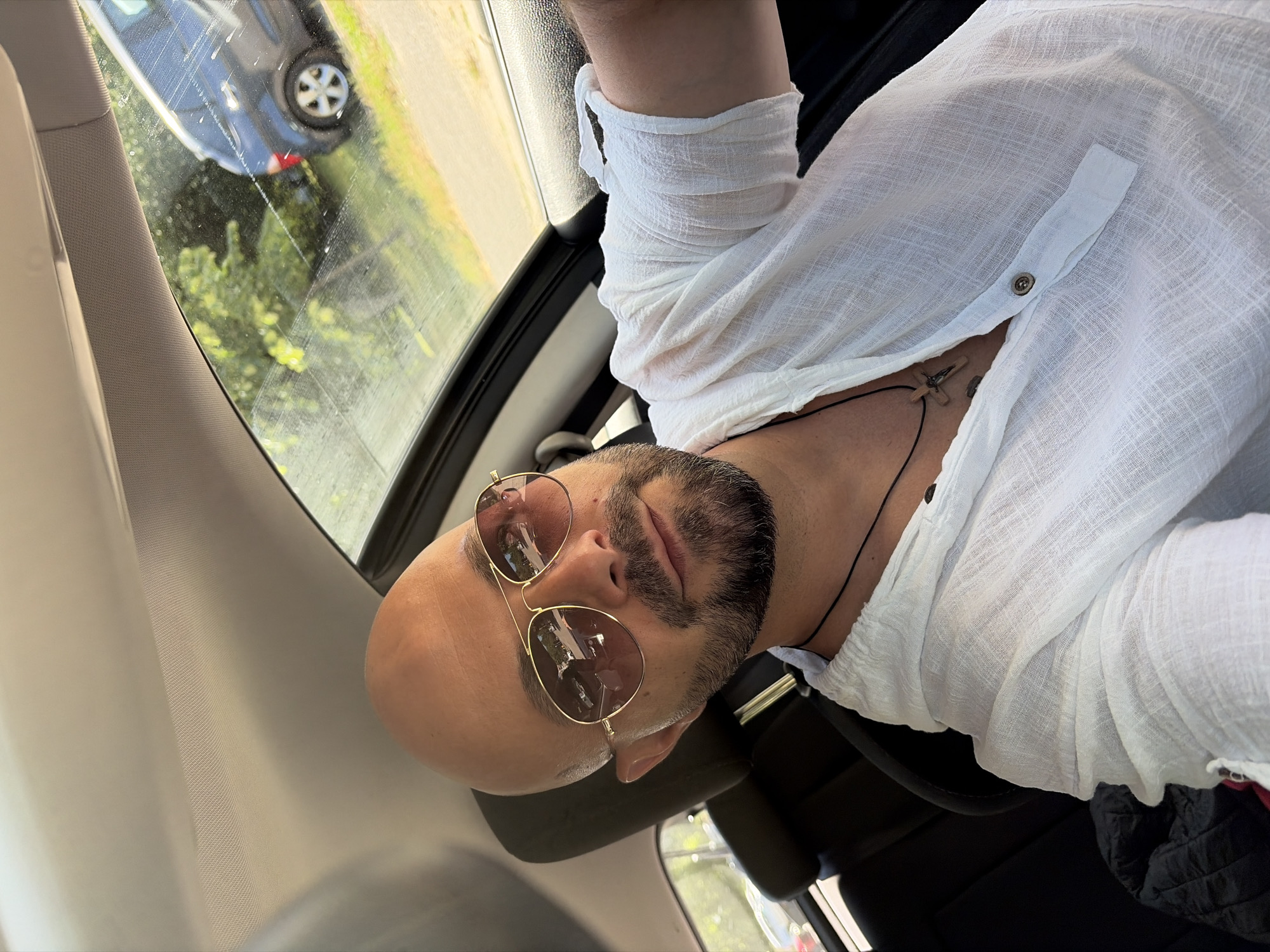 The image shows a man with a shaved head and a beard wearing sunglasses and a white shirt, sitting in the driver's seat of a car. He is looking at the camera, and his right arm is resting on the car door. Outside the car, trees and another car are visible. The scene appears to be in a natural environment, possibly a park or a rural area, given the greenery and the sunlit trees. The image does not contain any specific visual cues to suggest a location within the city of Sofia, Bulgaria.