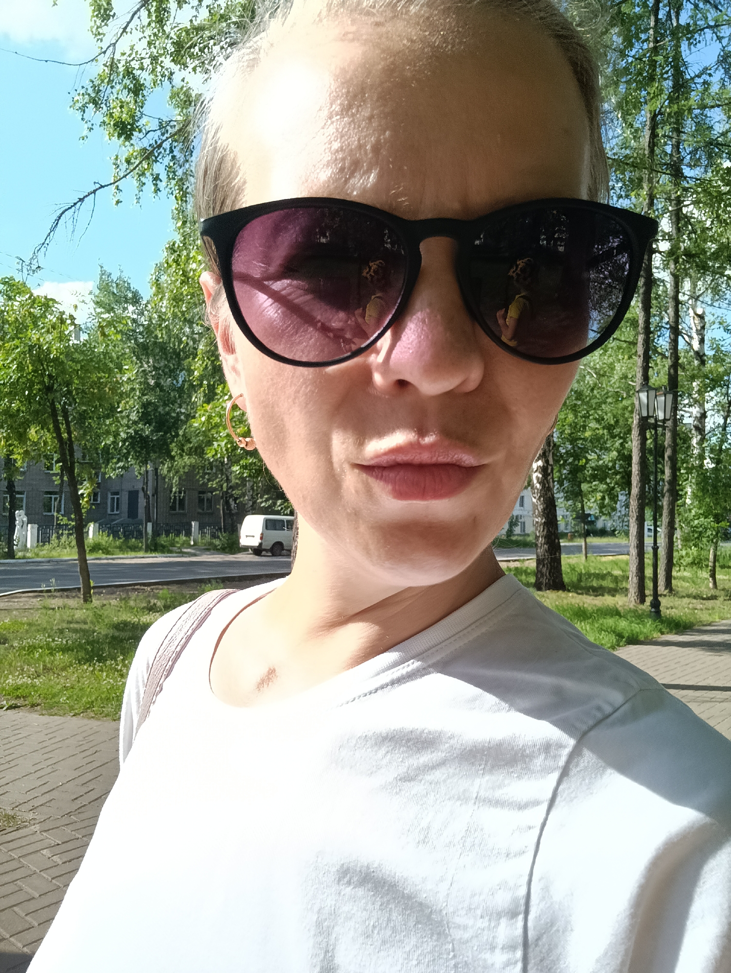 This is a selfie taken outdoors on a sunny day. The subject is a person wearing sunglasses and a white t-shirt, with a light-colored strap from a bag visible on their shoulder. The background shows trees, buildings, a road, a sidewalk, and a white van parked on the side of the road. The scene appears to be in a residential or park area. The lighting suggests it is daytime. The person's expression is neutral. There is no visible text in the image.