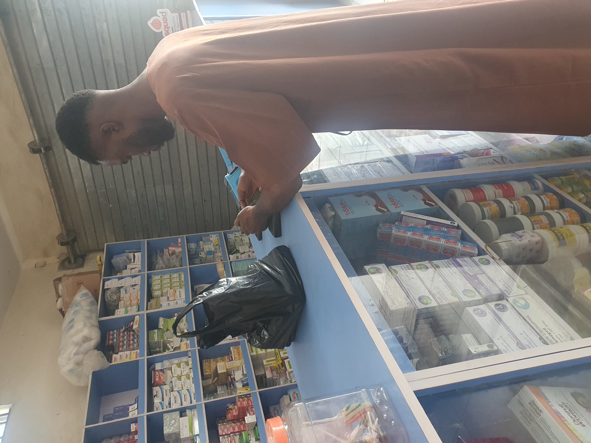 A man with a beard, wearing an orange shirt, is at a pharmacy in Gwagwalada, Nigeria. He is leaning over a glass display counter filled with various medications. A black plastic bag is on the counter next to him. Shelves behind the counter are stocked with additional pharmaceutical products. Boxes on the shelves are labeled with names like "IBUCAP" and "Combigack." Medications on the counter are named "Lisinopril" and "Celoxigen."